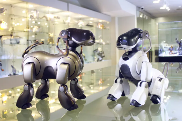 Twee robot-honden, een zilveren en een witte, tentoongesteld in een glazen vitrine met andere robots op de achtergrond.
