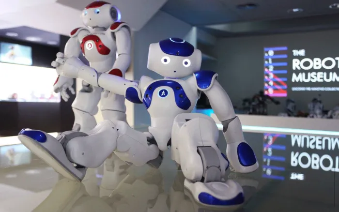 Twee wit-blauwe humanoïde robots op een reflecterend oppervlak met op de achtergrond een bord met de tekst THE ROBOT MUSEUM