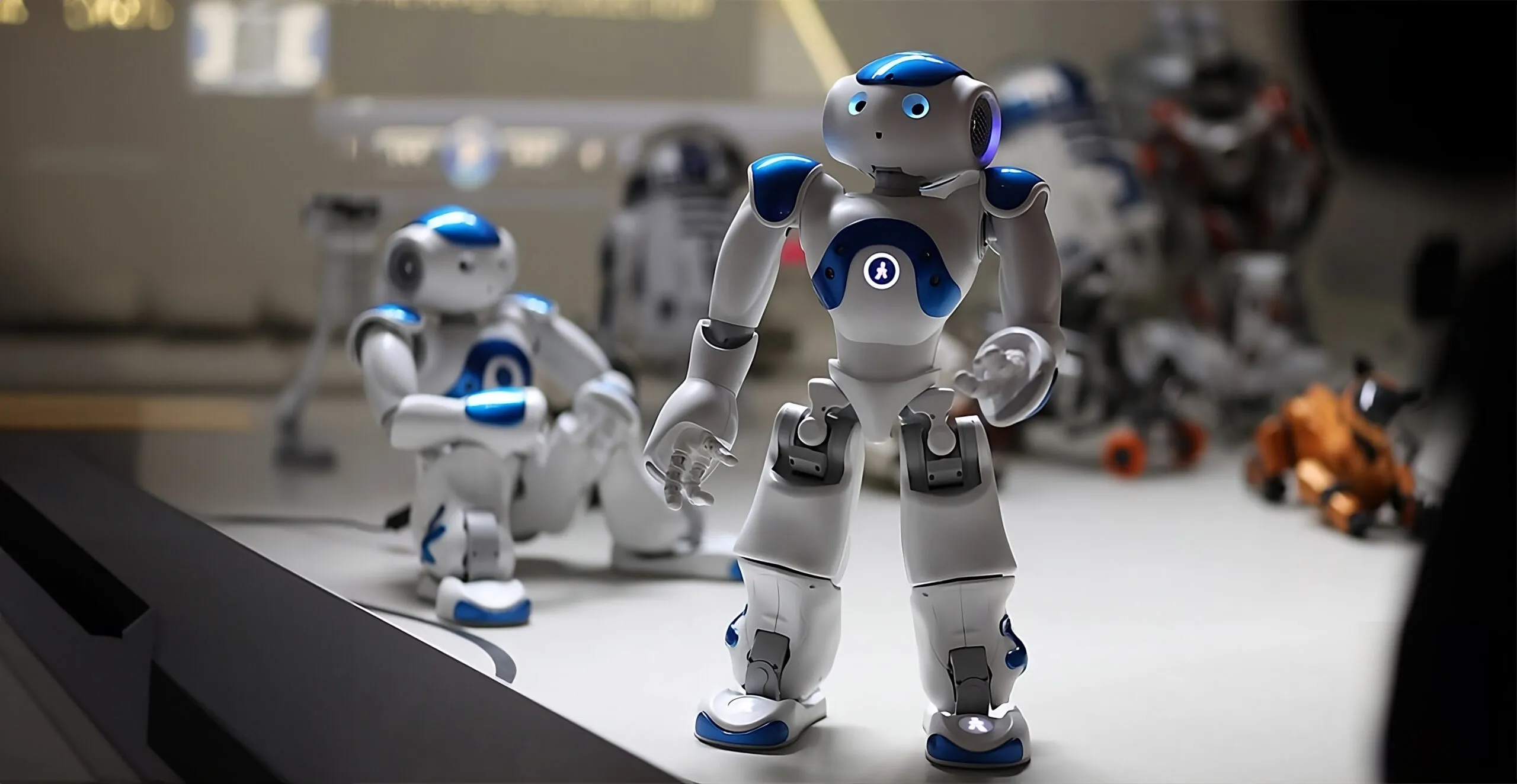Een witte en blauwe humanoïde robot met een blauw hoofd en borstpaneel staat op een displaytafel, met andere robotfiguren op de achtergrond.
