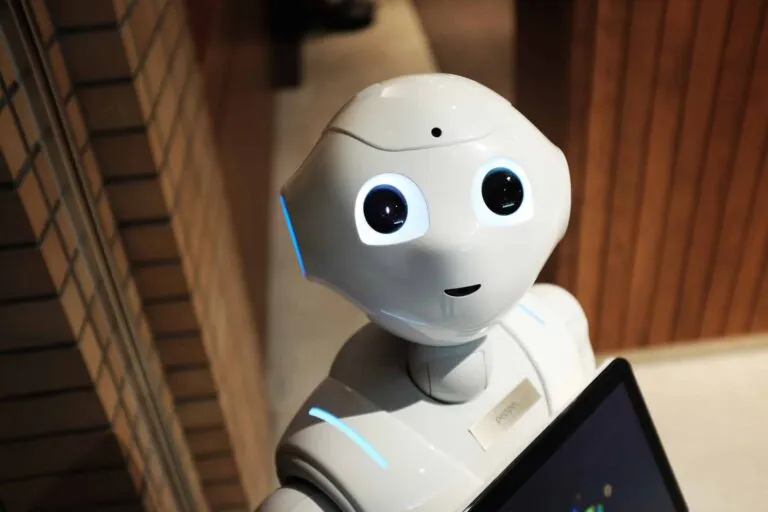 Een witte humanoïde robot met gloeiende blauwe ogen en accenten, staand binnenshuis nabij een bakstenen muur.