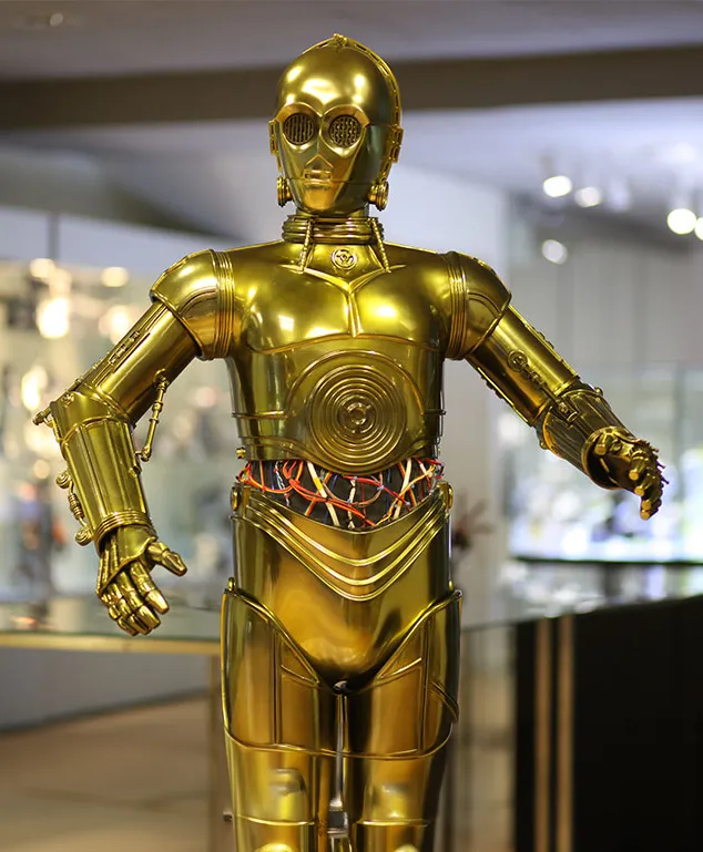 Een gouden humanoïde robot met een circulair borstpaneel en gelede ledematen tentoongesteld in een museum