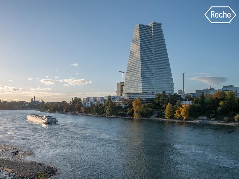 Tall glass skyscraper Roche Tower rising above the Rhine river with a passenger boat passing by and autumn trees along the bank
