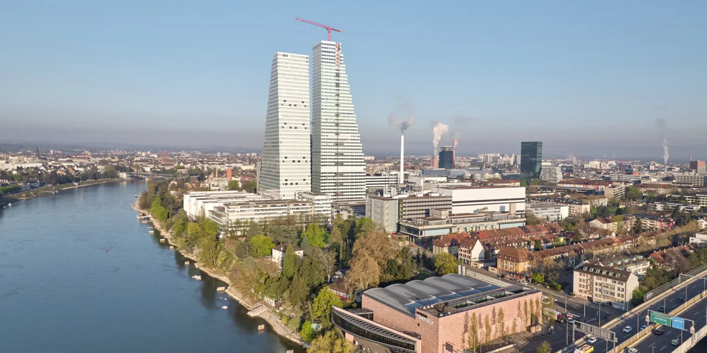 Aerial view of the twin Roche towers rising above Basel's cityscape along the Rhine river, with a construction crane atop one tower