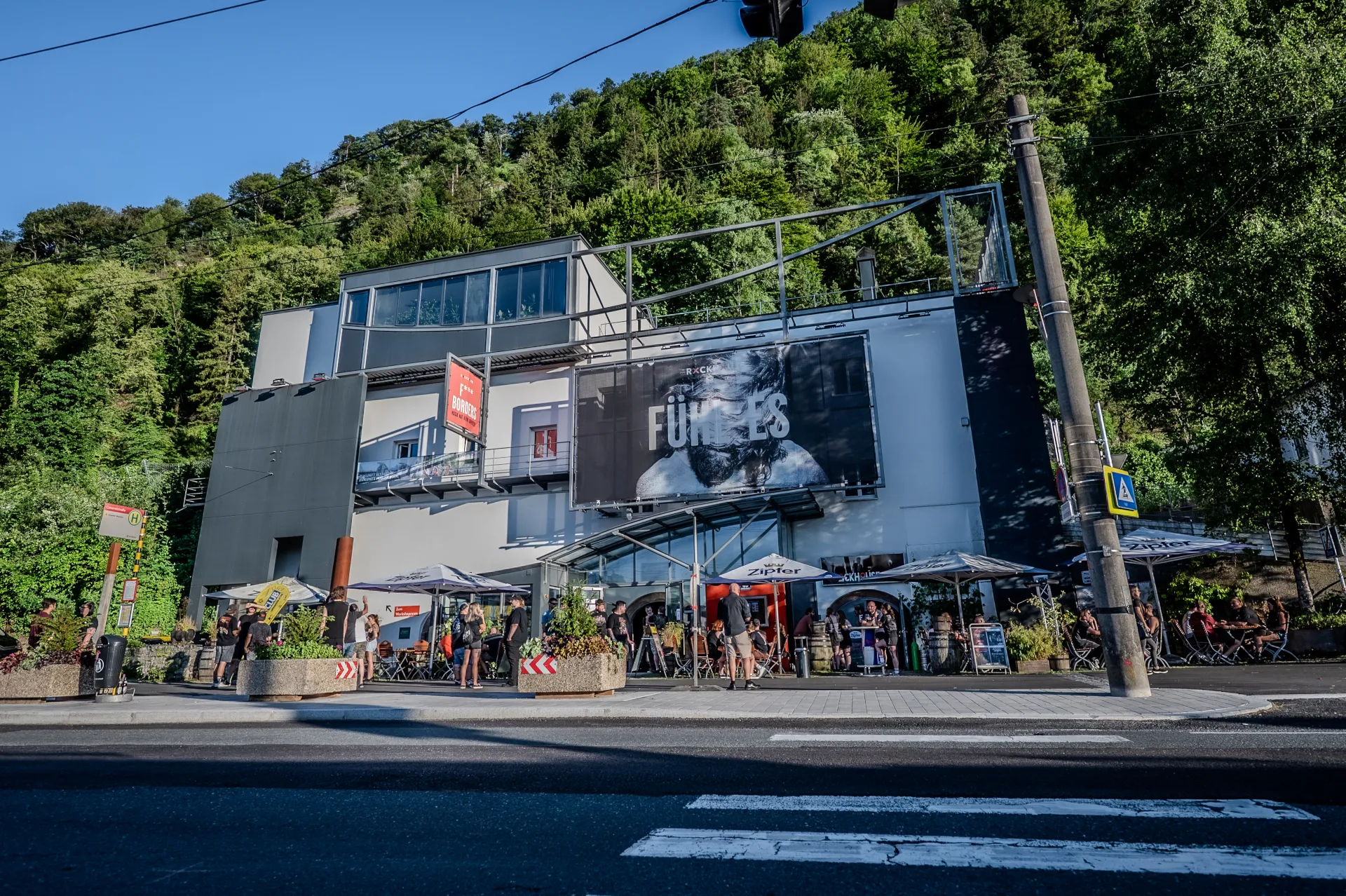 Buitenaanzicht van Rockhouse Salzburg met een moderne gevel, een groot concertspandoek, een terras met parasols en een beboste heuvel op de achtergrond.