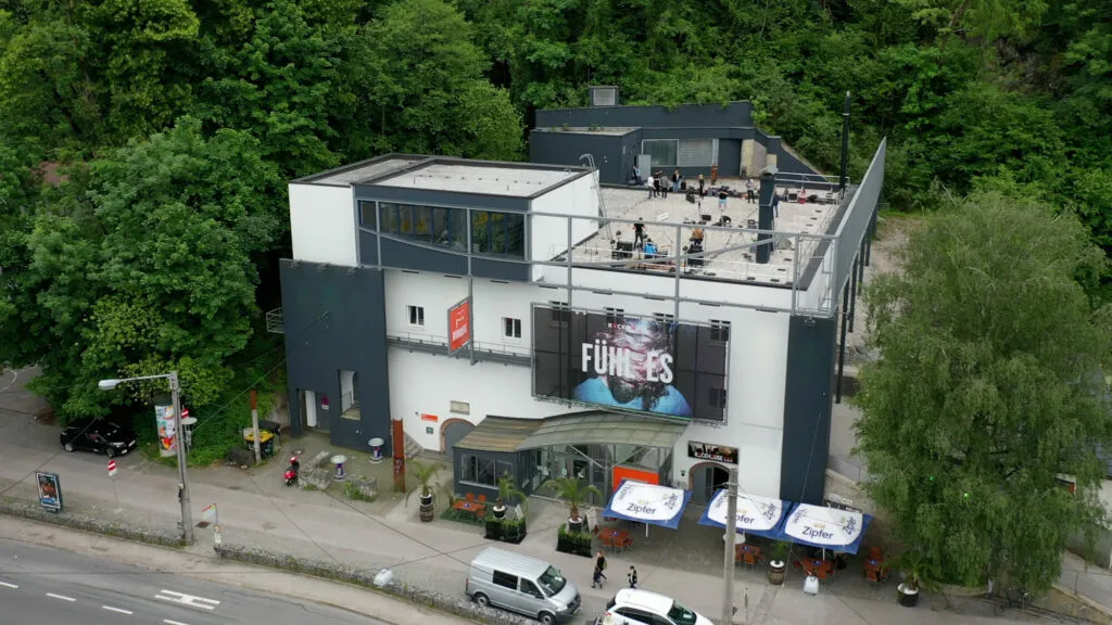 Luchtfoto van een modern wit en donkergrijs gebouw met een grote FUNKIES-banner, omringd door weelderige groene bomen, met een straat en geparkeerde voertuigen beneden.