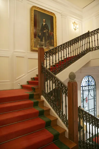 Schody z czerwonym dywanem i zieloną lamówką, ozdobnymi czarnymi żelaznymi balustradami oraz dużym oprawionym portretem na białej, boazeryjnej ścianie.