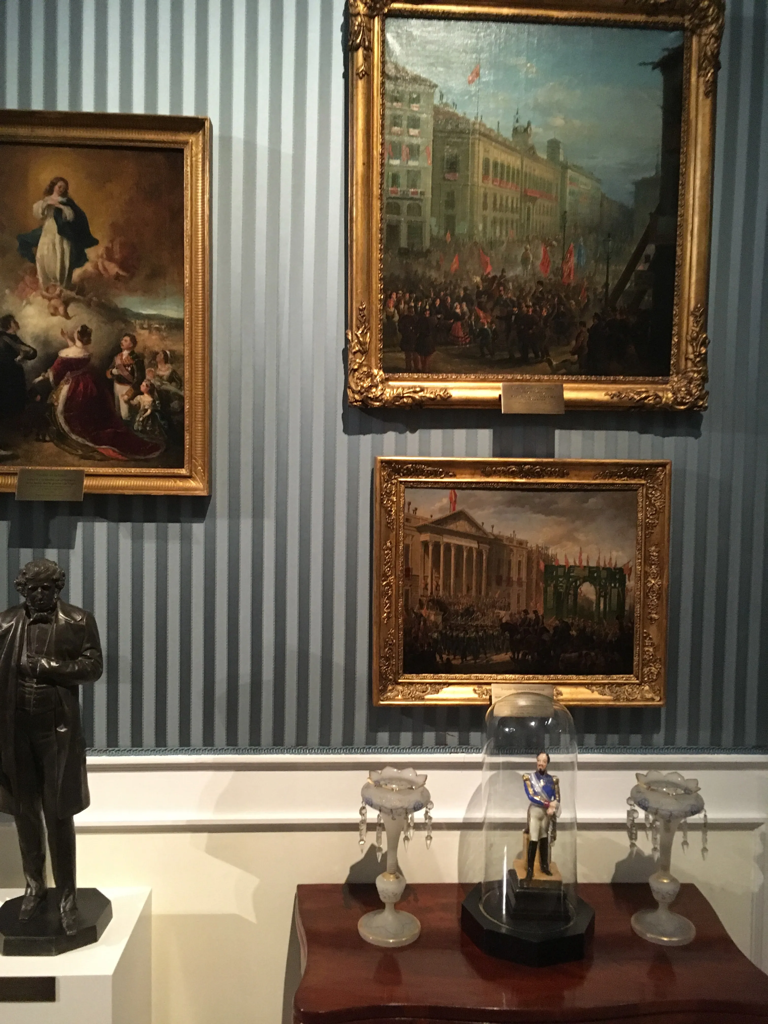 Pinturas en marcos dorados, una estatua y una figura bajo una cúpula de cristal sobre una mesa de madera contra una pared azul a rayas