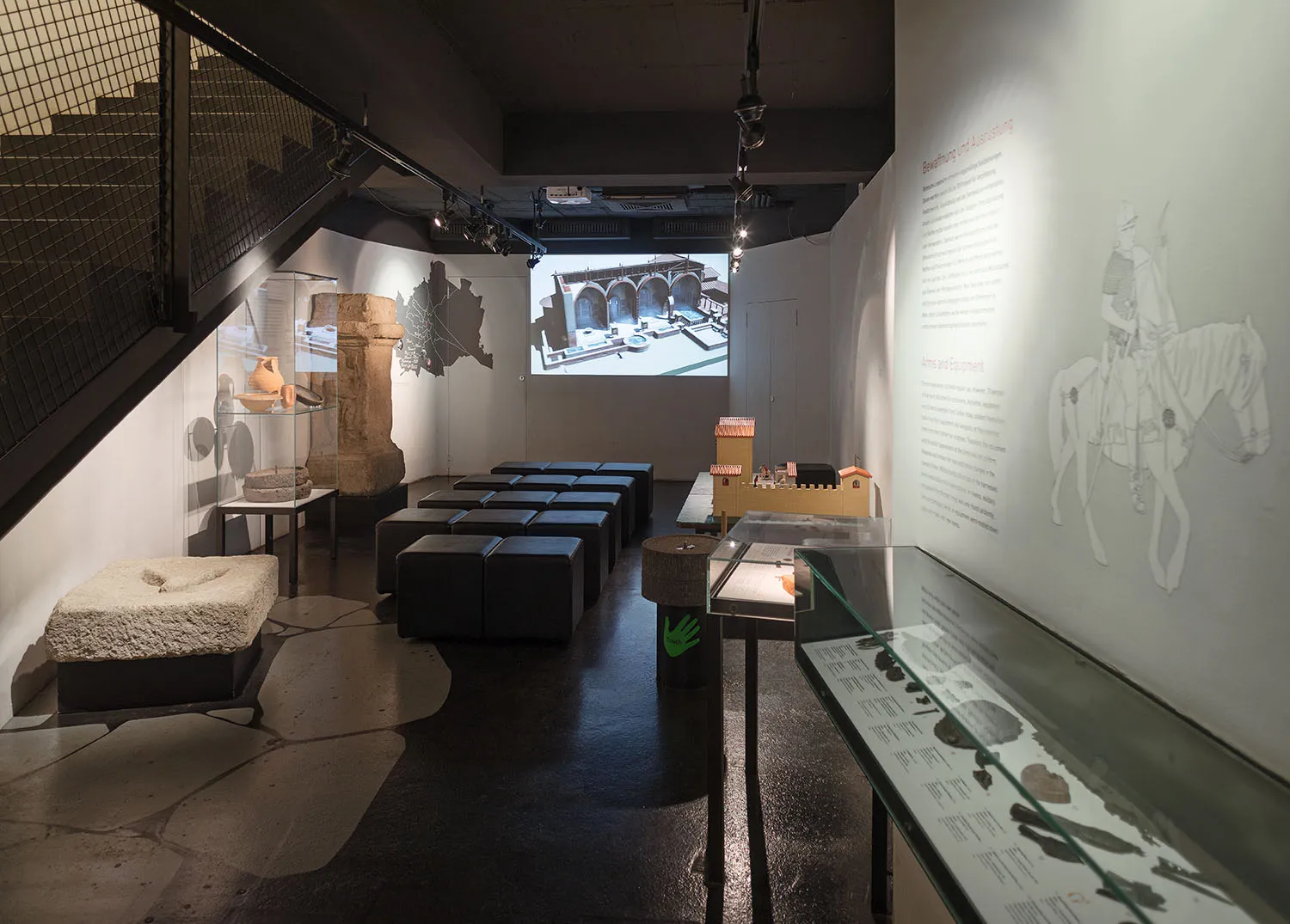 Wnętrze Römermuseum z artefaktami w gablotach, ekranem projekcyjnym i muralem przedstawiającym rzymskiego żołnierza