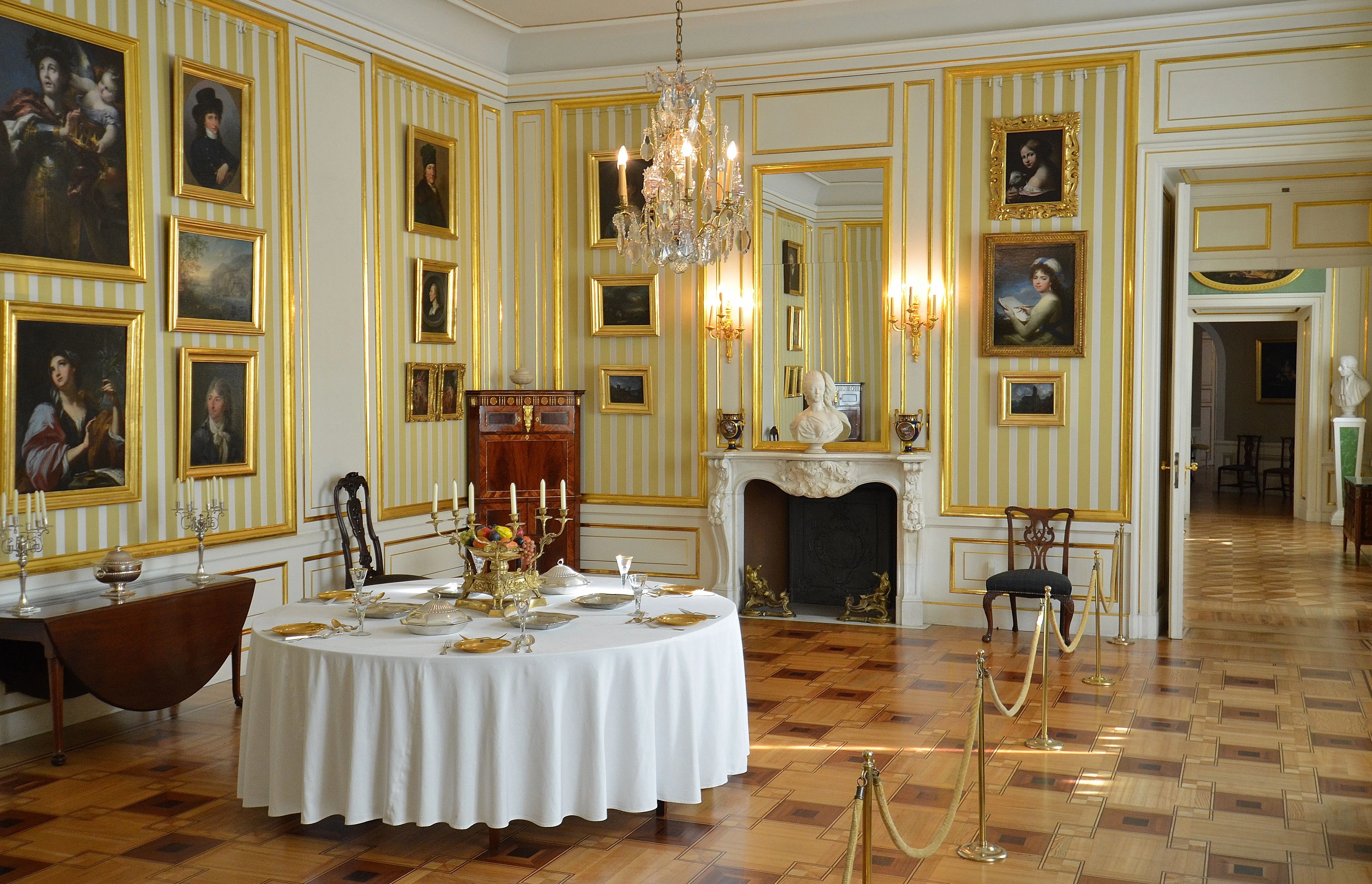 Sierlijke kamer met geel-wit gestreepte muren, een kroonluchter, een open haard en ingelijste schilderijen, met een ronde tafel bedekt met een wit tafelkleed en gedekt met borden en kaarsen.