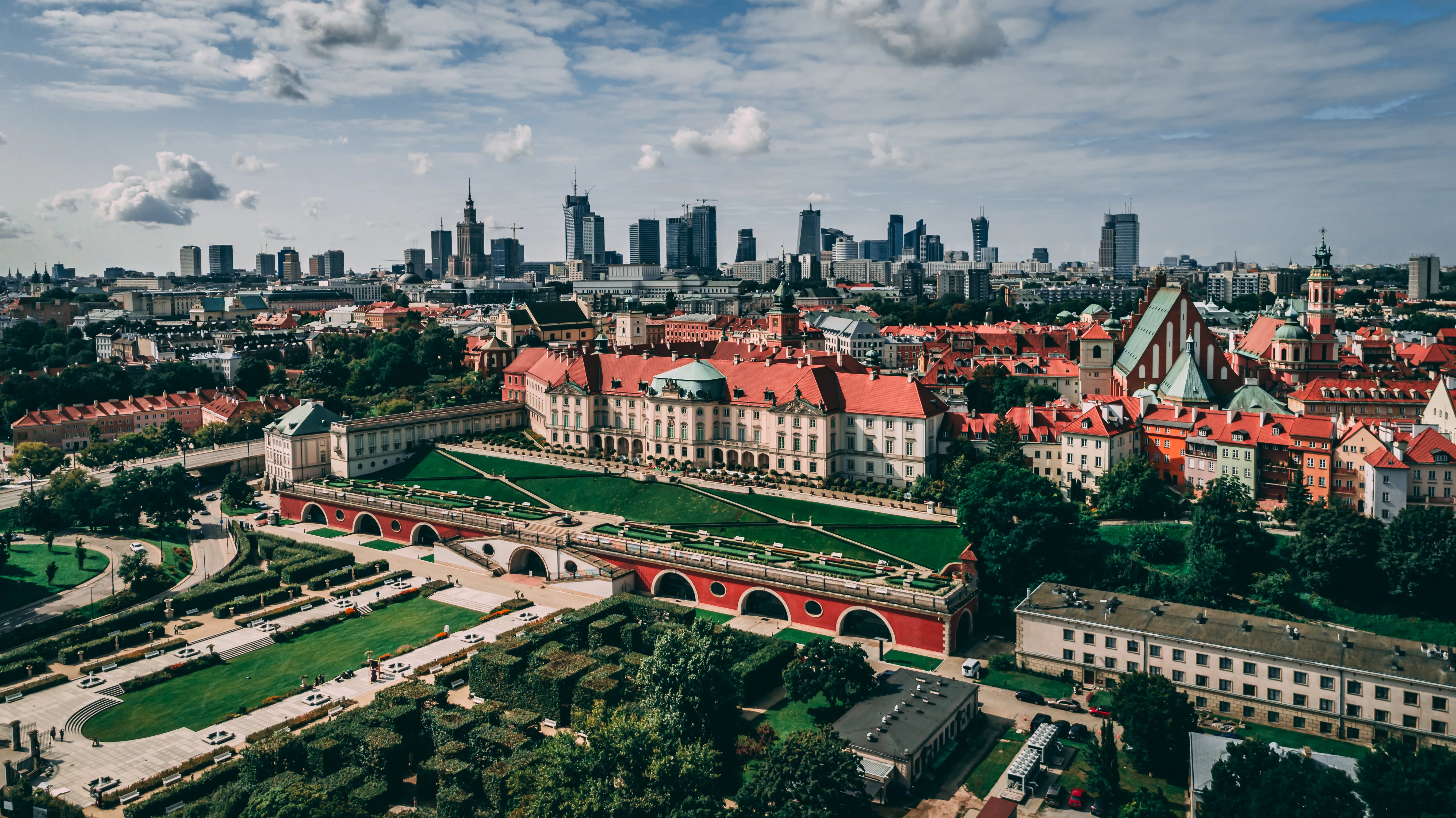 Widok z lotu ptaka na Zamek Królewski w Warszawie z czerwonymi dachami budynków i zielonymi ogrodami, na tle nowoczesnej panoramy miasta