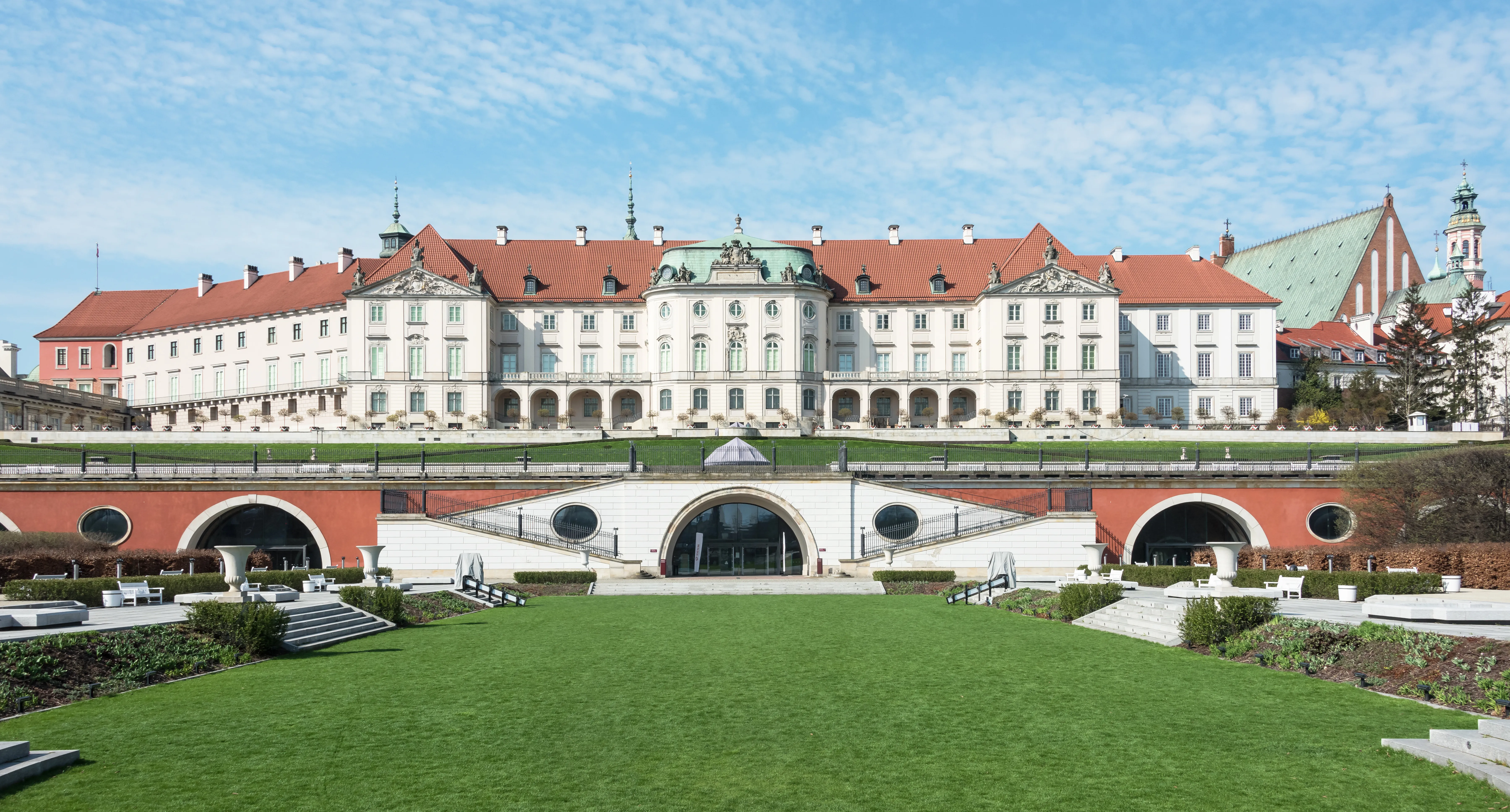 Zamek Królewski w Warszawie z czerwonym, ceramicznym dachem i białą fasadą, widziany z zielonego terenu z mostem i łukami na pierwszym planie pod błękitnym niebem.