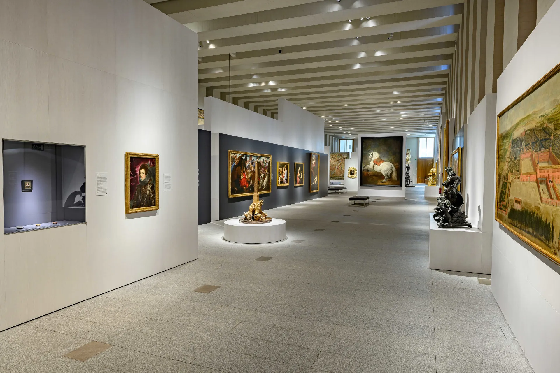 Długa galeria z obrazami na białych ścianach, centralną ekspozycją rzeźb i dużym gobelinem na końcu.