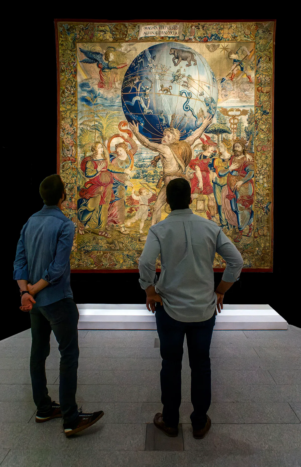 Dwie osoby oglądające duży gobelin z centralną postacią trzymającą kulę ziemską, otoczoną postaciami w galerii