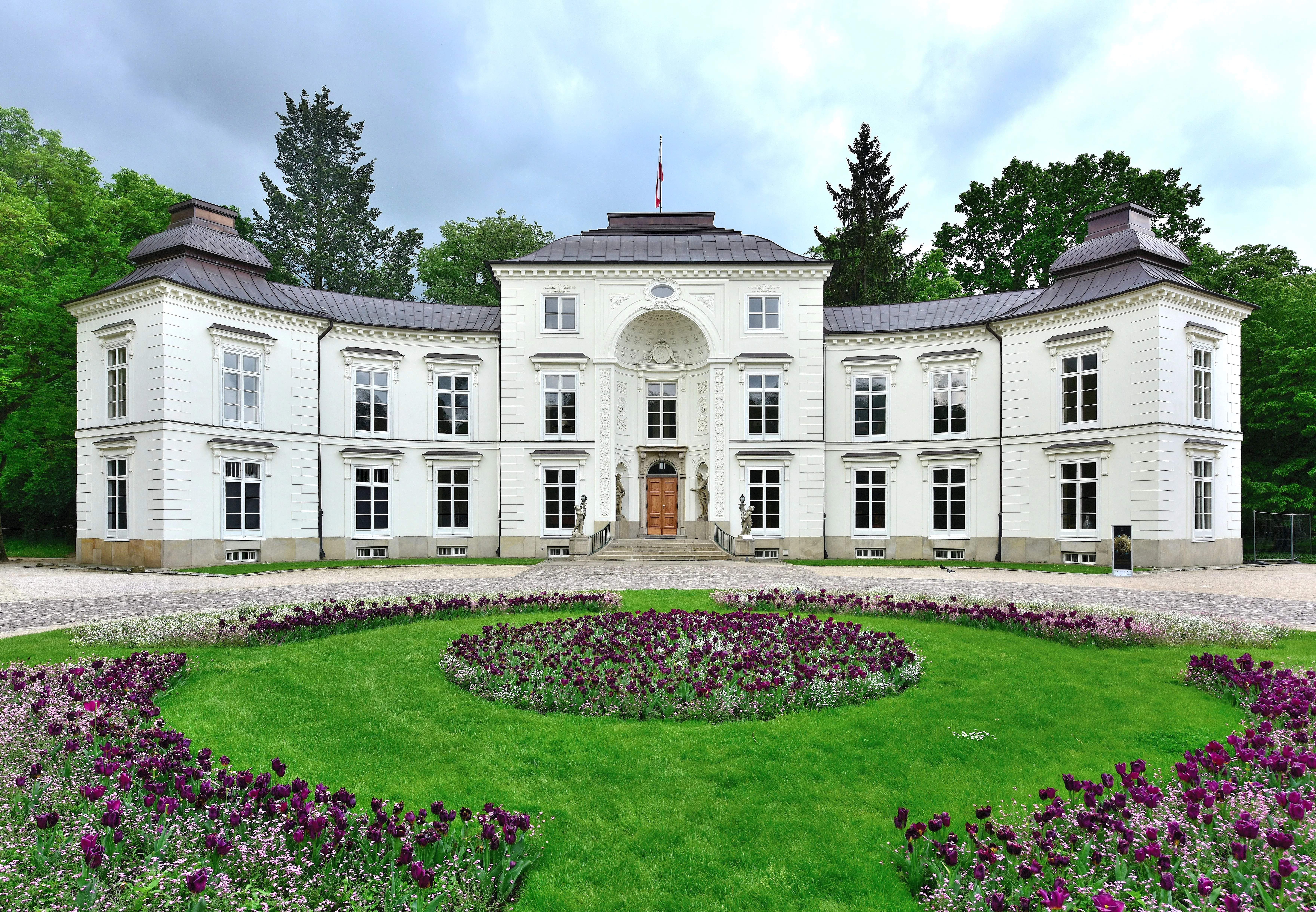 White neoclassical palace with symmetrical windows and a central entrance, surrounded by green lawns and purple flowers.