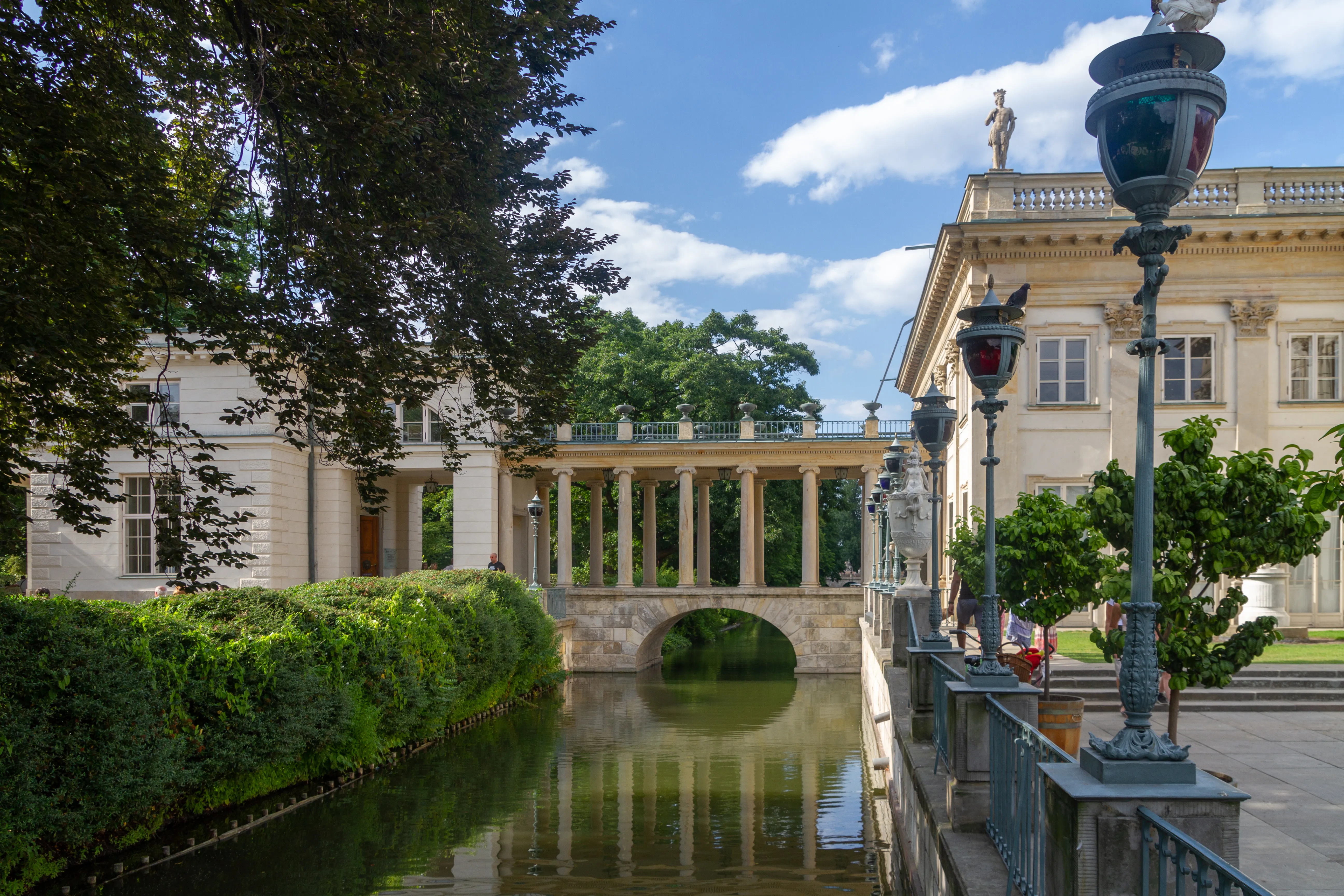 Classical pavilions and a stone bridge over a canal in a park with green hedges and trees under a blue sky