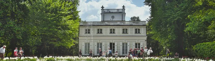 Wit neoklassiek paleis met een centrale koepel en zuilen, omgeven door groene bomen en witte bloemen, met mensen op de voorgrond