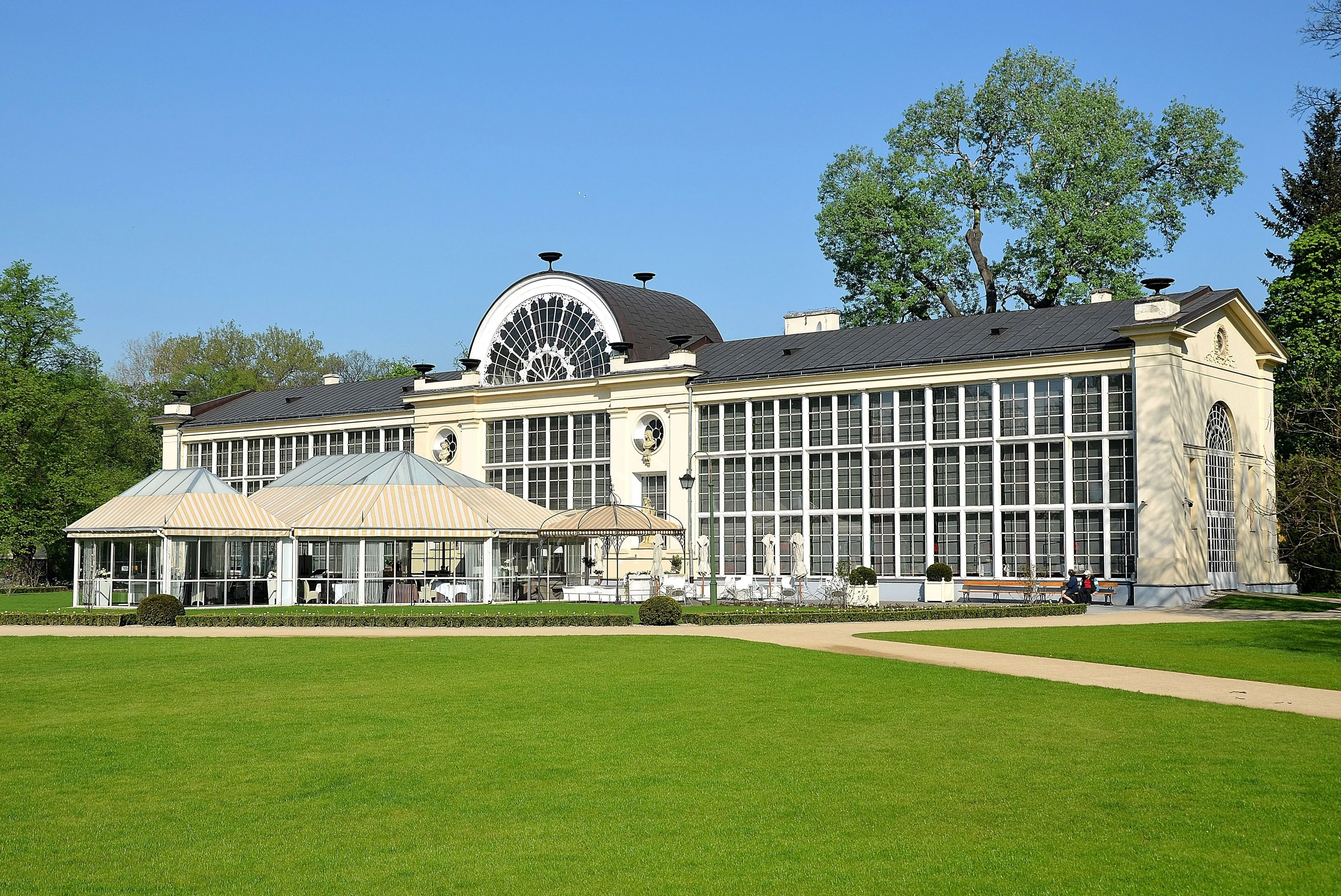 Neoclassicistisch gebouw met grote glazen ramen en een gebogen dak, omgeven door groene gazons en bomen onder een helderblauwe lucht