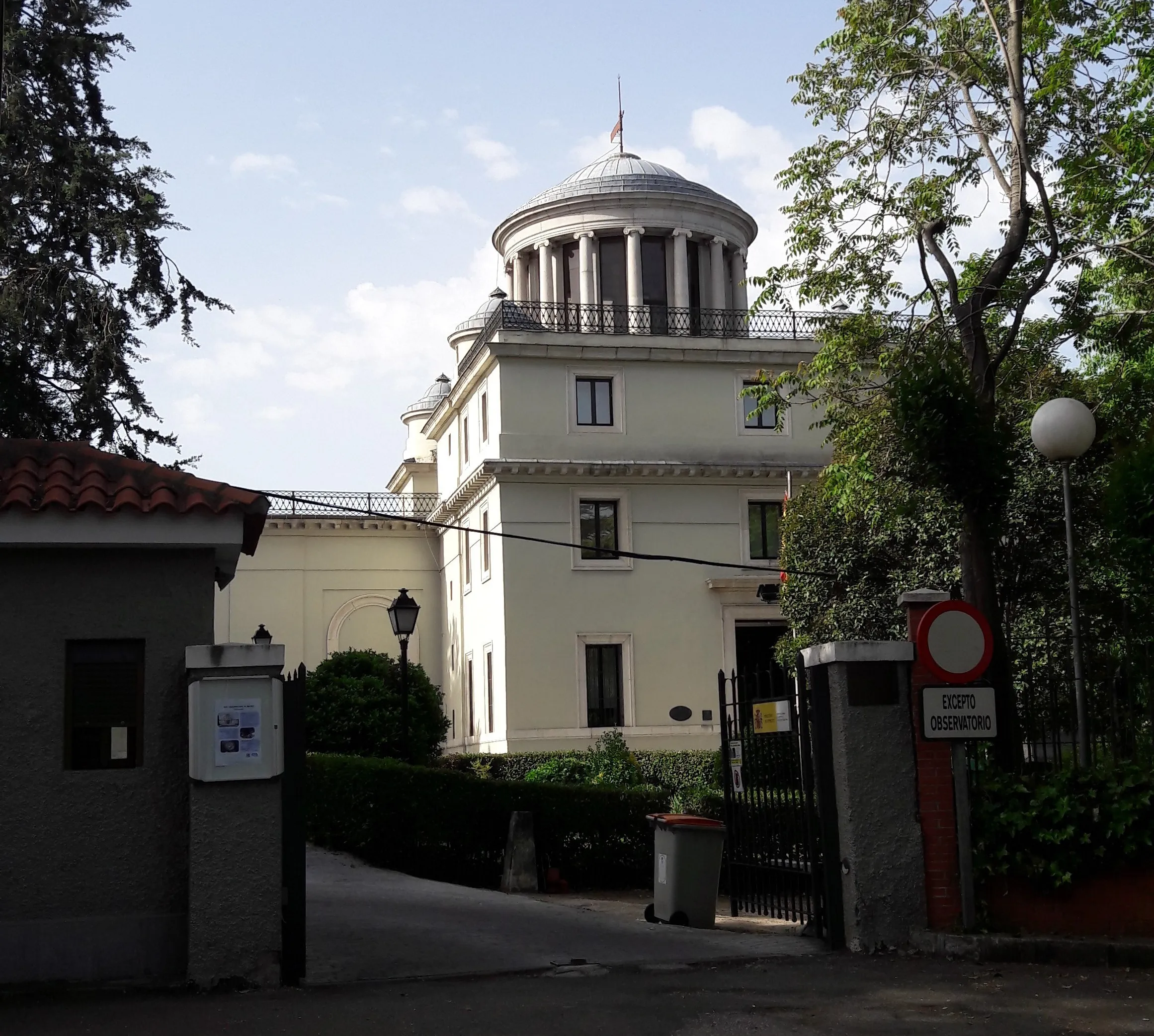 Biały neoklasycystyczny budynek z kopułą i kolumnami, otoczony drzewami i bramą wejściową