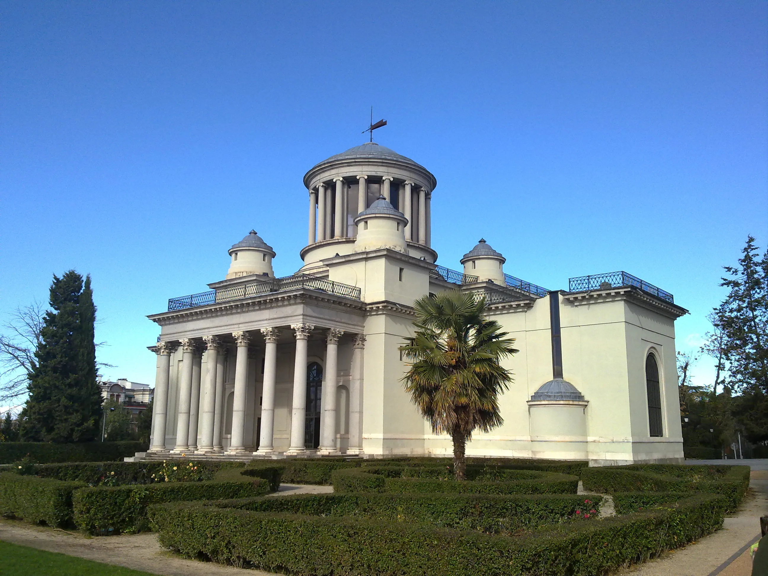 Neoklasycystyczny budynek z centralną kopułą i kolumnami, otoczony zielonymi żywopłotami i drzewami pod czystym, błękitnym niebem