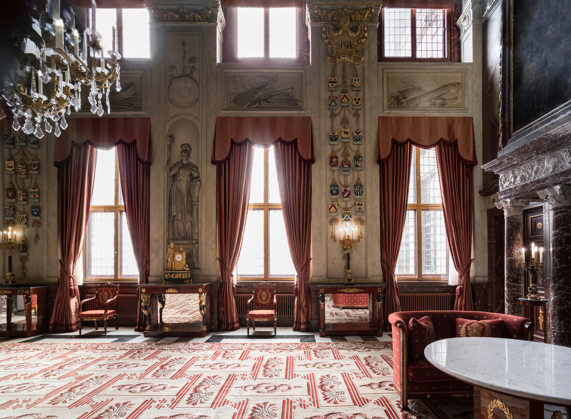 Spacious royal hall with tall windows, red drapery, ornate chandeliers, and intricate red-and-white patterned carpet