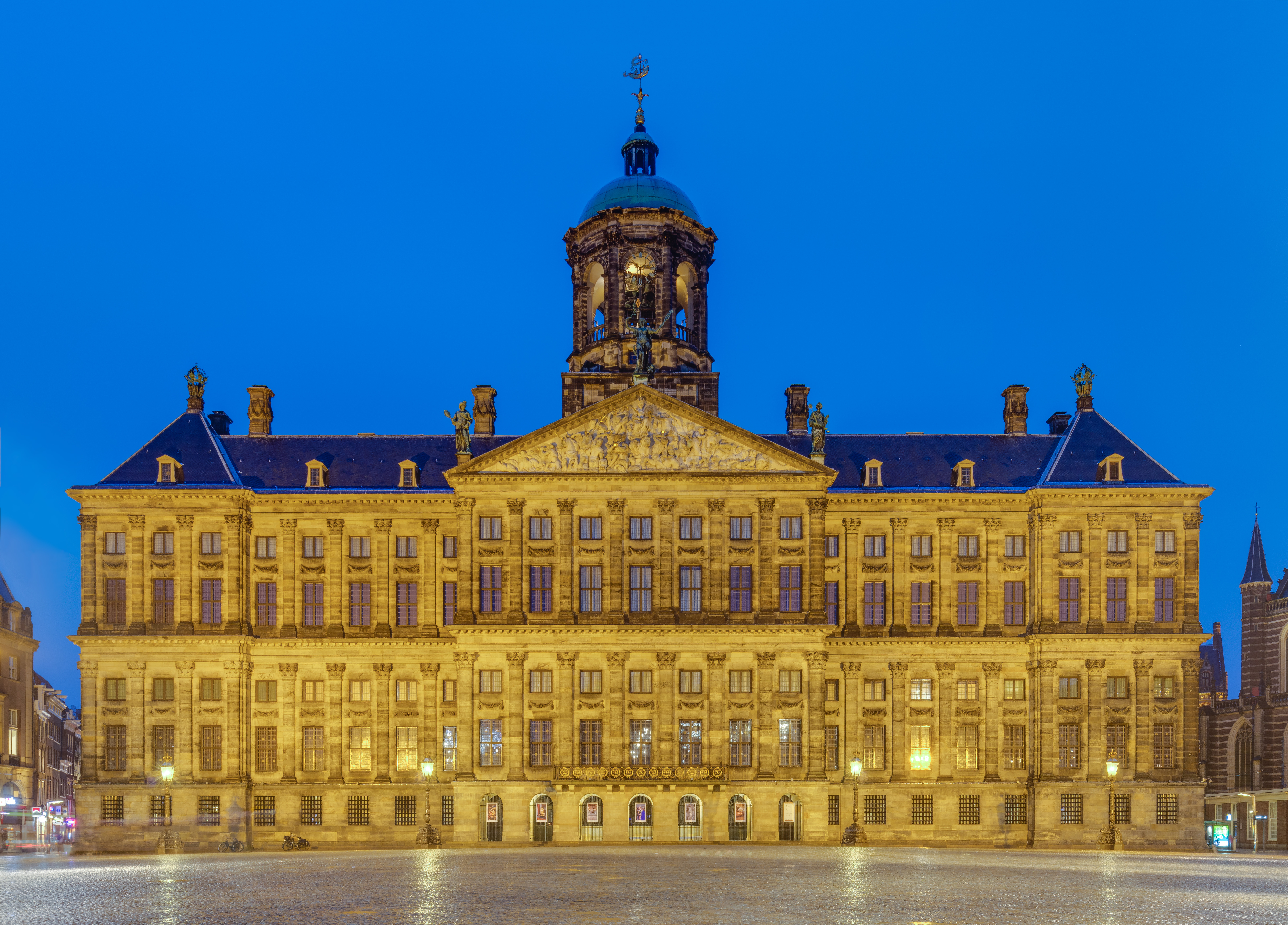 El Palacio Real de Ámsterdam por la noche, un gran edificio iluminado con una torre de reloj central y fachada simétrica bajo un cielo azul oscuro