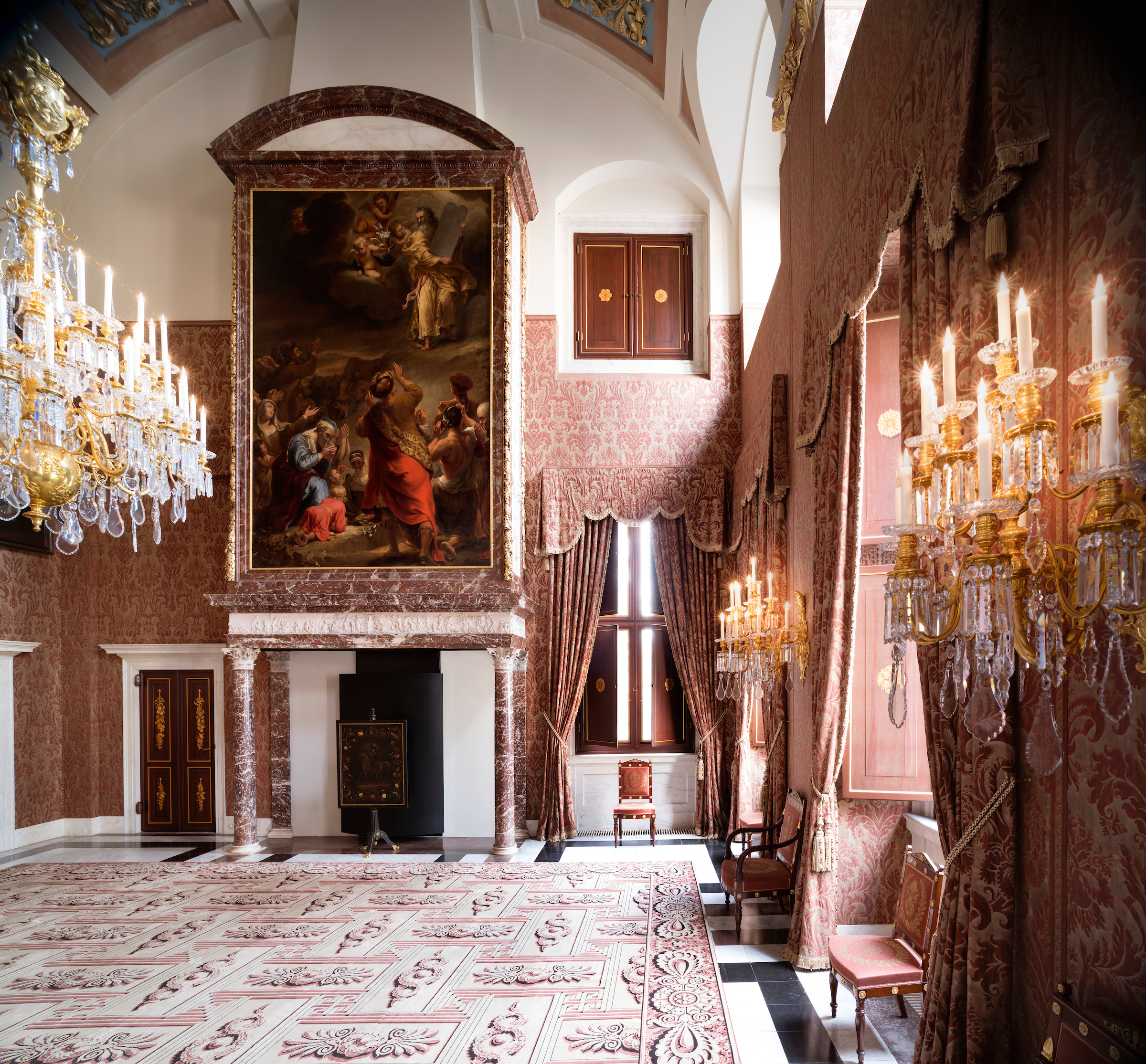 Gran sala interior del Palacio Real de Ámsterdam con candelabros ornamentados, un gran cuadro histórico, suelo de mármol estampado y cortinas de terciopelo rojo