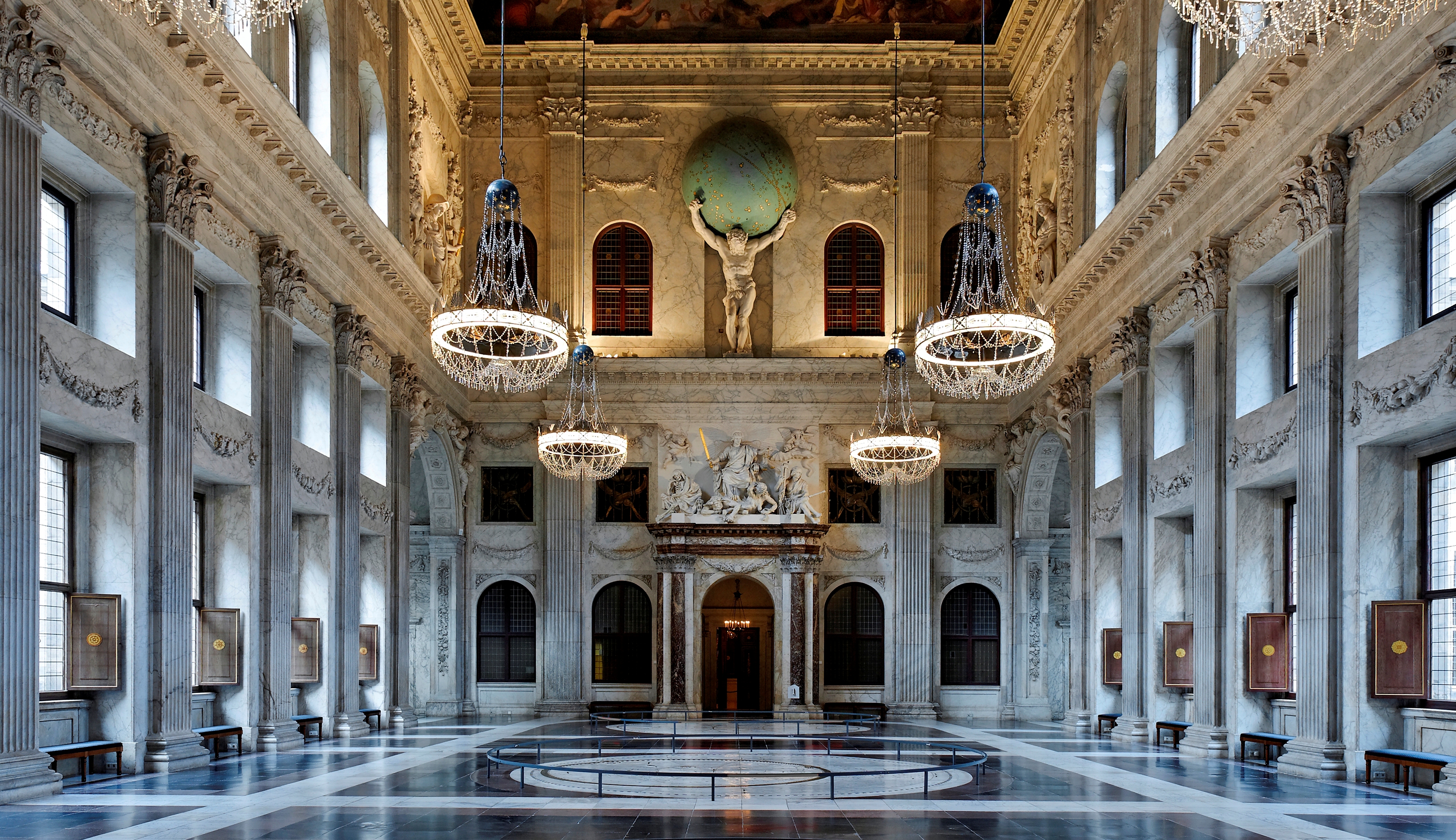 Gran salón con altas columnas, candelabros ornamentados, una estatua central y un suelo de mármol reflectante en un palacio histórico.