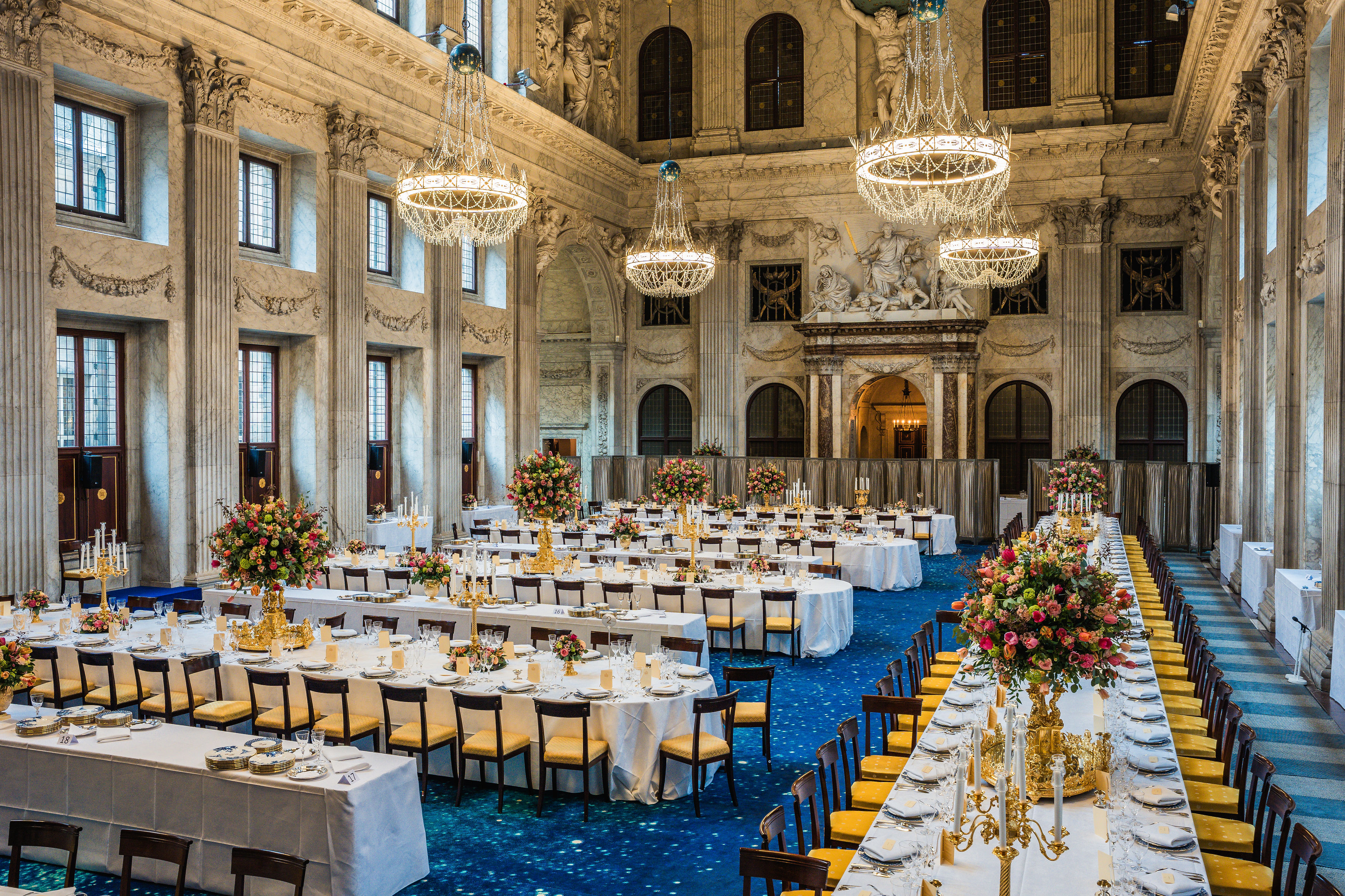 Gran salón del Palacio Real de Ámsterdam con alfombra azul estampada, mesas puestas con arreglos florales, candelabros dorados y lámparas de araña de cristal bajo altos techos ornamentados.