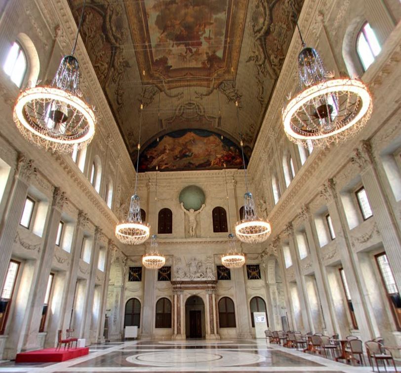 Interior del gran salón del Palacio Real de Ámsterdam con frescos ornamentados en el techo, grandes candelabros y hileras de ventanas