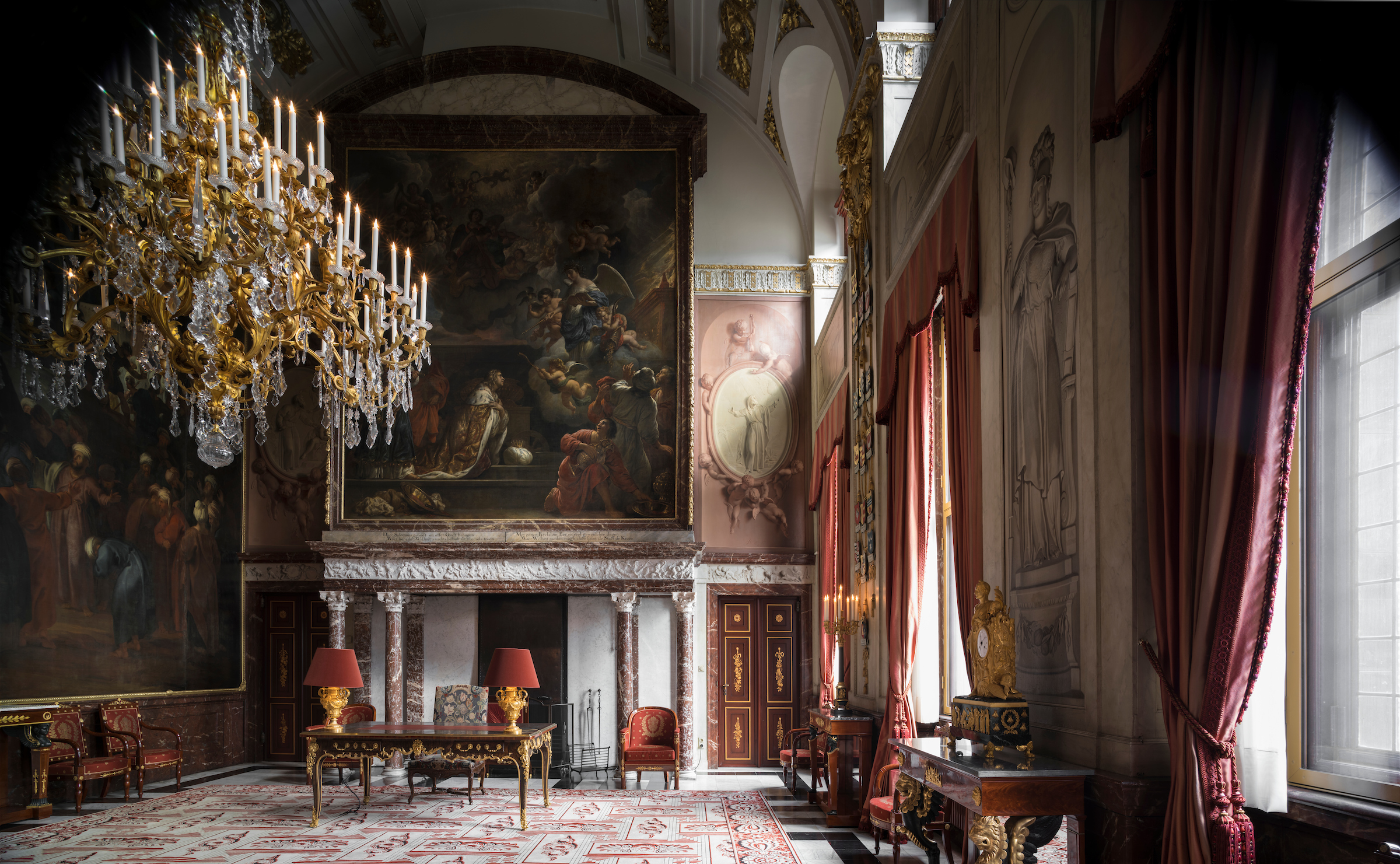 Interior del gran salón con grandes candelabros de cristal, pinturas históricas al óleo, cortinas de terciopelo rojo, muebles ornamentados y chimenea de mármol en el Palacio Real de Ámsterdam