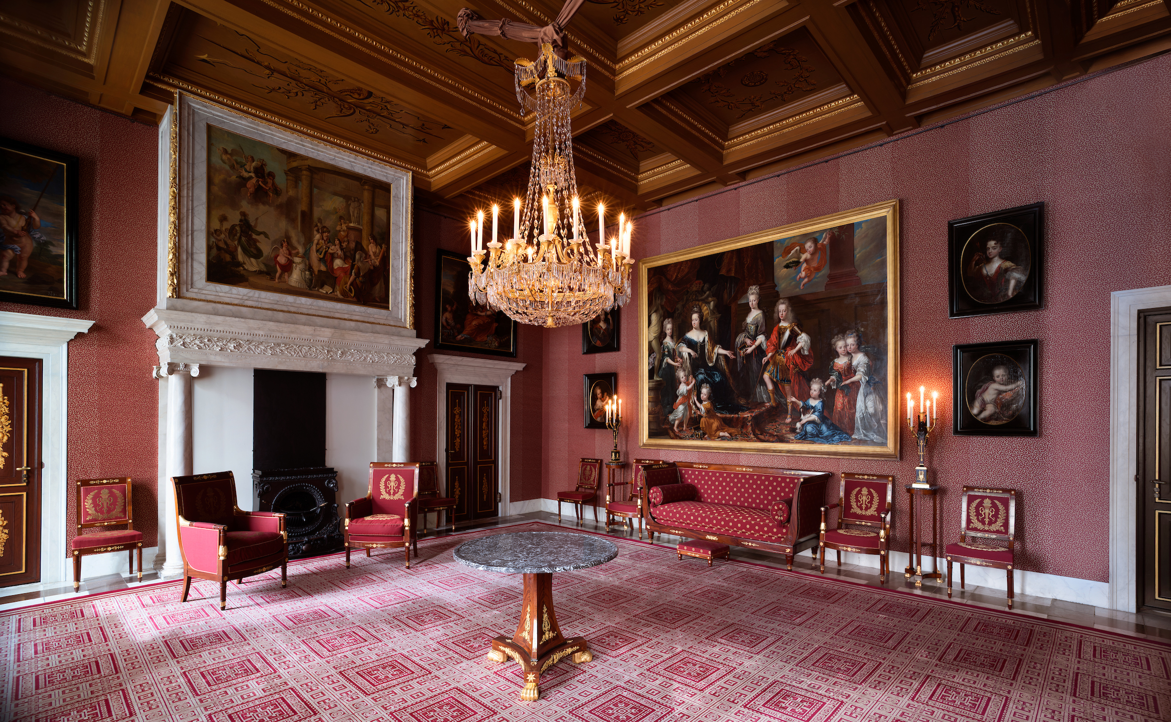 Gran salón con paredes rosas, candelabro ornamentado, grandes cuadros enmarcados, sillas tapizadas rojas y alfombra estampada