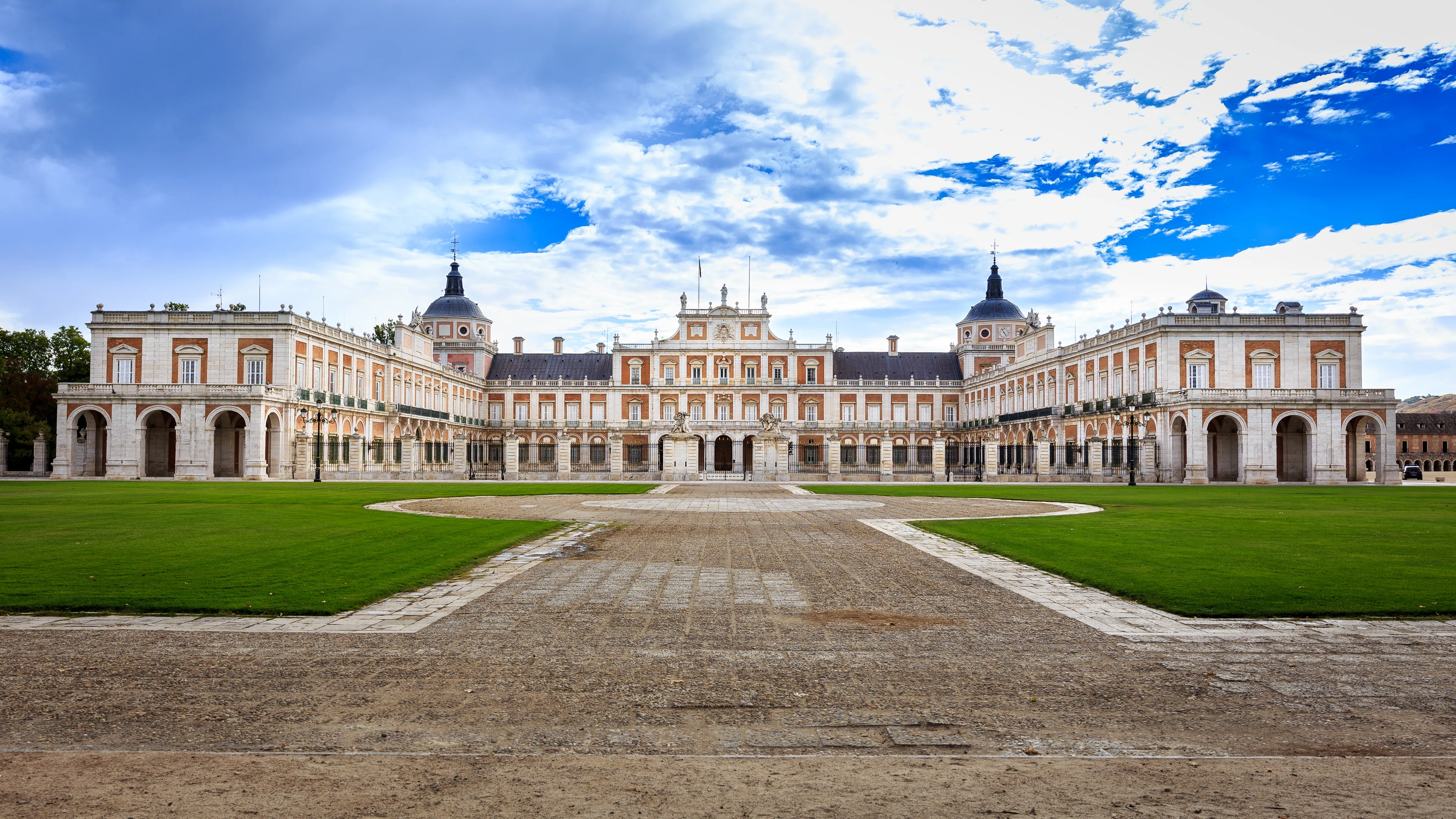 Gran palacio barroco con dos cúpulas y una fachada simétrica, rodeado de céspedes verdes y un camino de piedra bajo un cielo azul