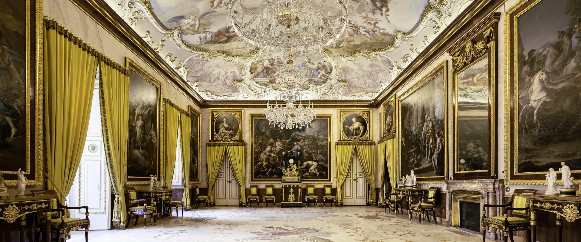 Ornato interior del Palacio Real de Aranjuez con cortinas doradas, una lámpara de araña y un techo con frescos