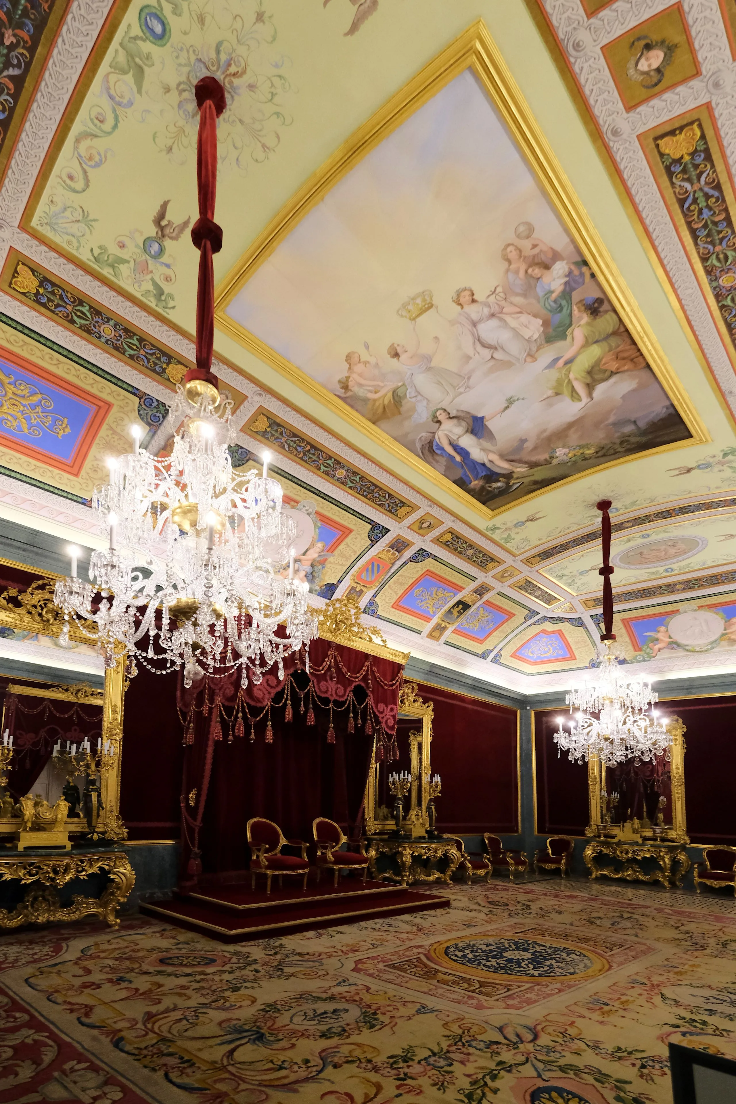 Interior ornamentado del Palacio Real de Aranjuez con una gran lámpara de araña, techo con frescos y un trono bajo un dosel rojo.