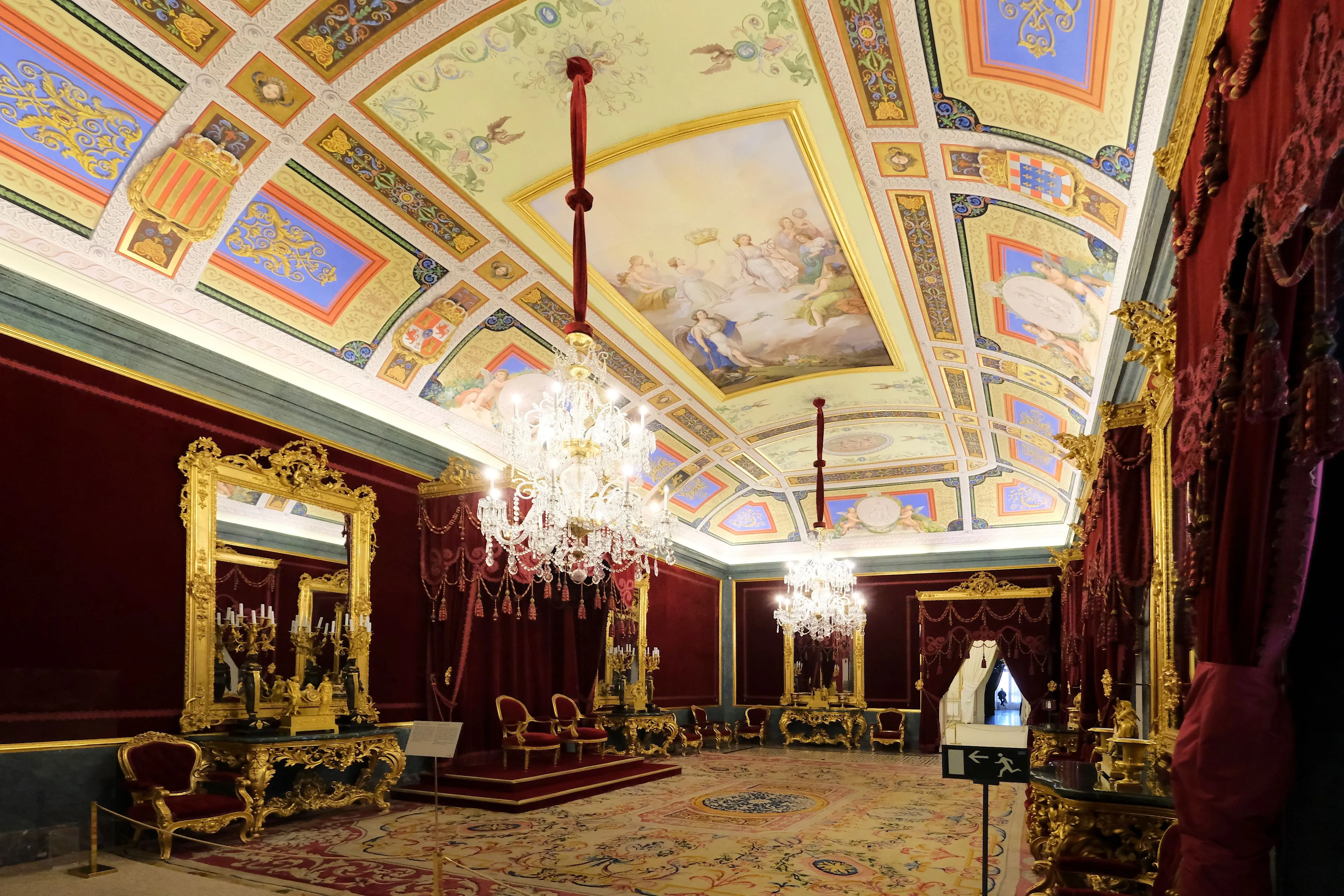 Interior ornamentado de una sala del trono real con muebles dorados, cortinas de terciopelo rojo, una gran lámpara de araña y un techo pintado con paneles decorativos