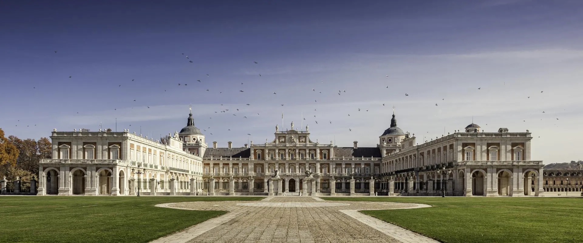 Gran palacio neoclásico con alas simétricas y cúpulas, rodeado de césped verde y un camino de piedra bajo un cielo azul claro