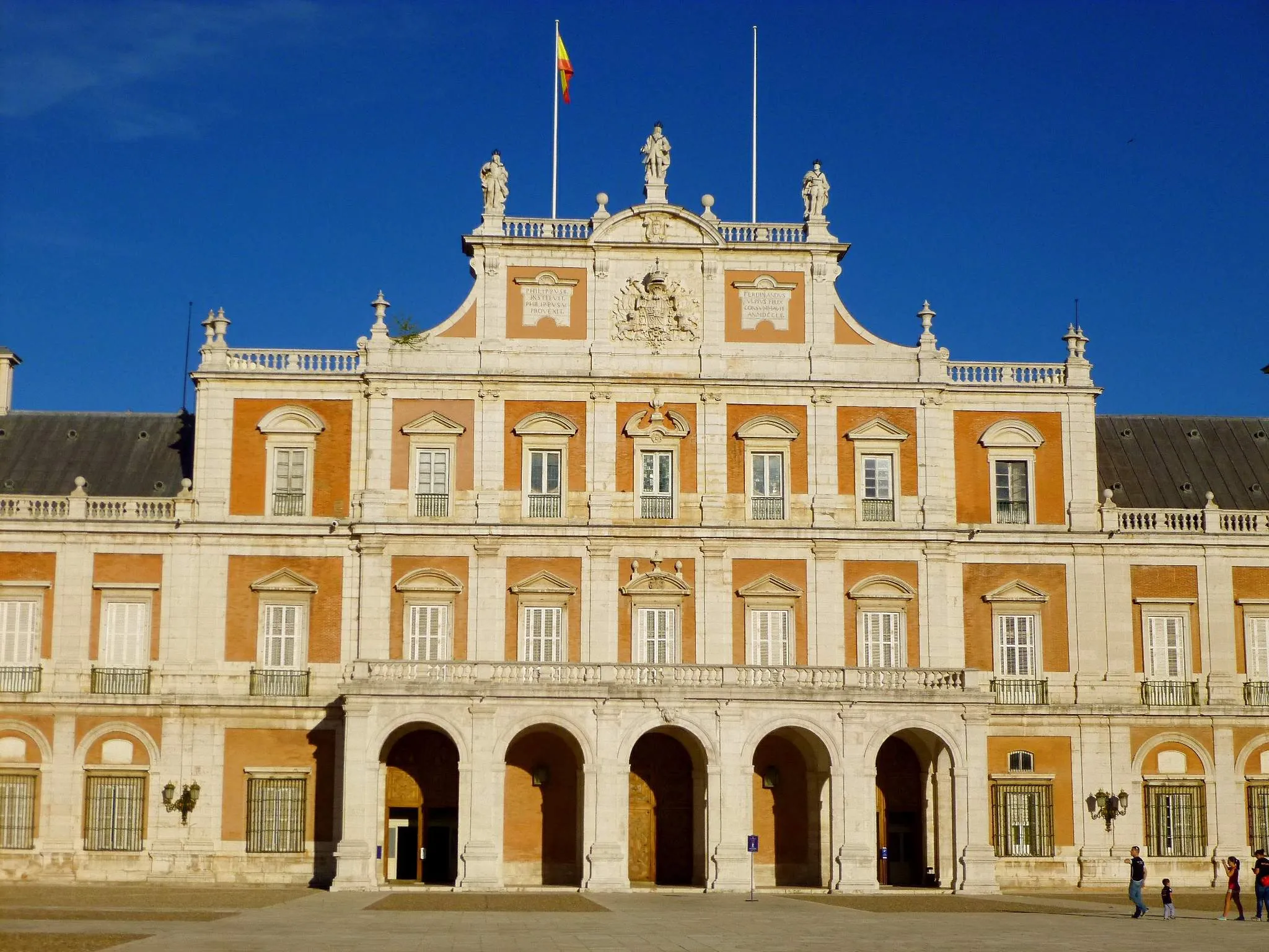 El Palacio Real de Aranjuez con su fachada ornamentada, entradas arqueadas y una bandera española ondeando en lo alto del edificio.