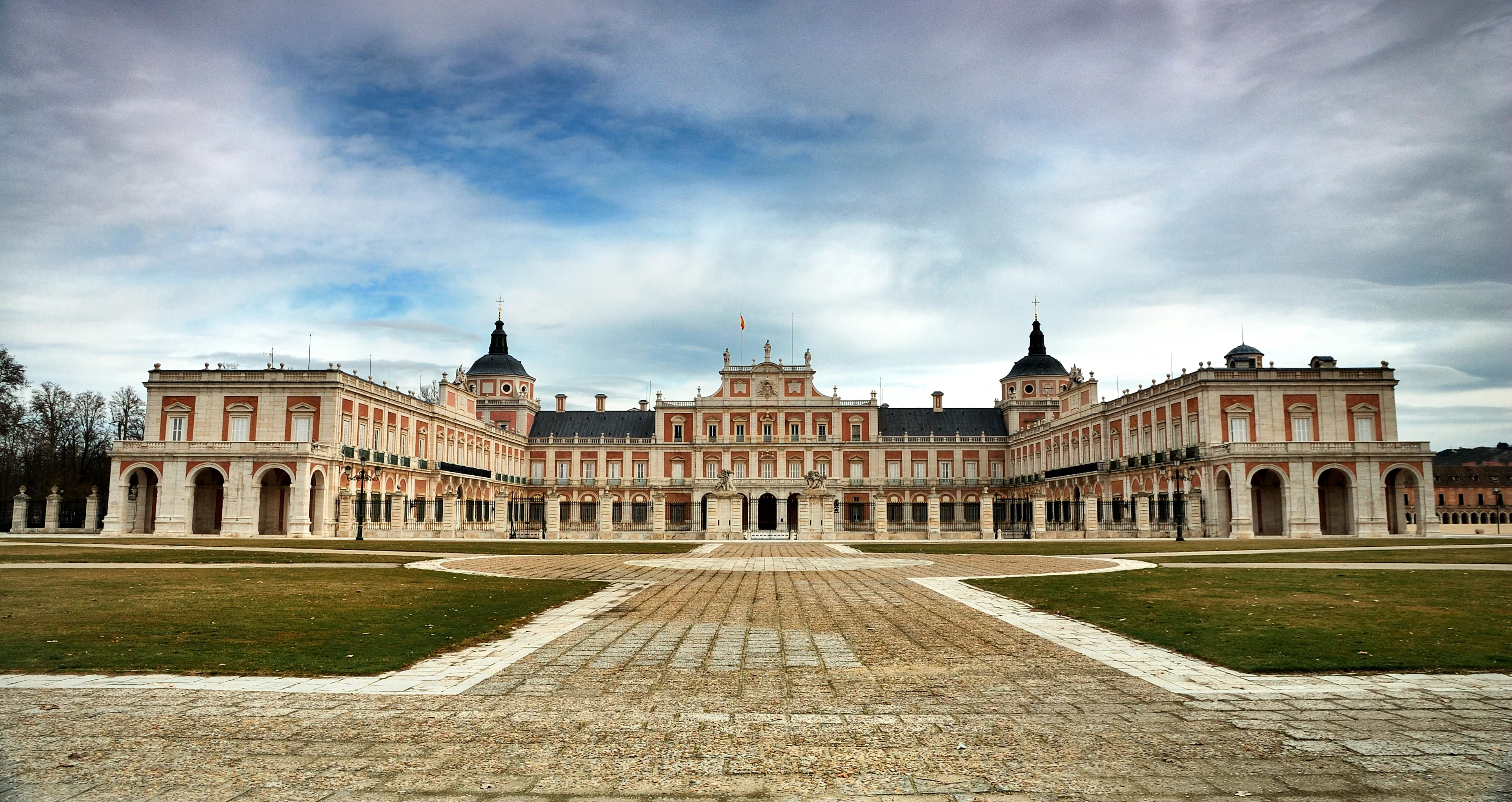 Una amplia vista del Palacio Real de Aranjuez, un gran edificio clásico con múltiples alas y torres, contra un cielo nublado, con un patio pavimentado y céspedes verdes en primer plano.