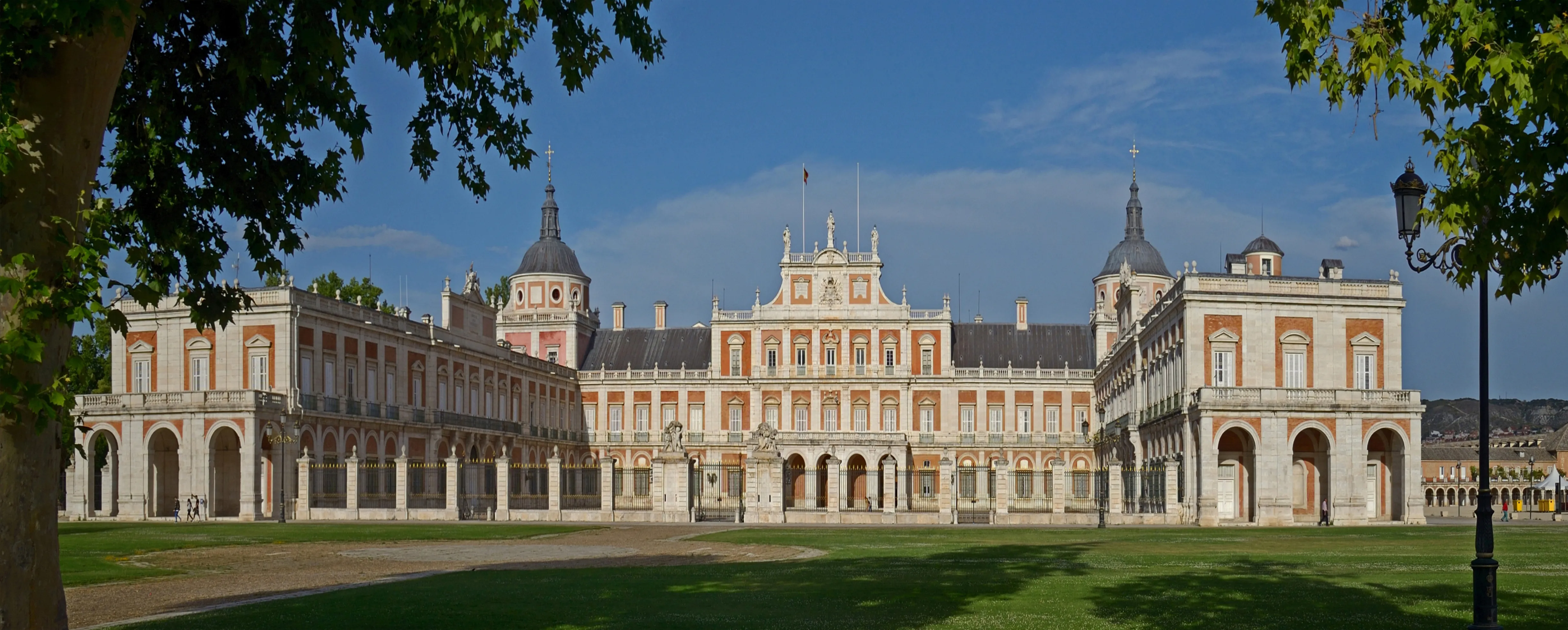 El Palacio Real de Aranjuez con su larga fachada, torres abovedadas y columnatas arqueadas, visto desde un césped verde bajo un cielo azul.