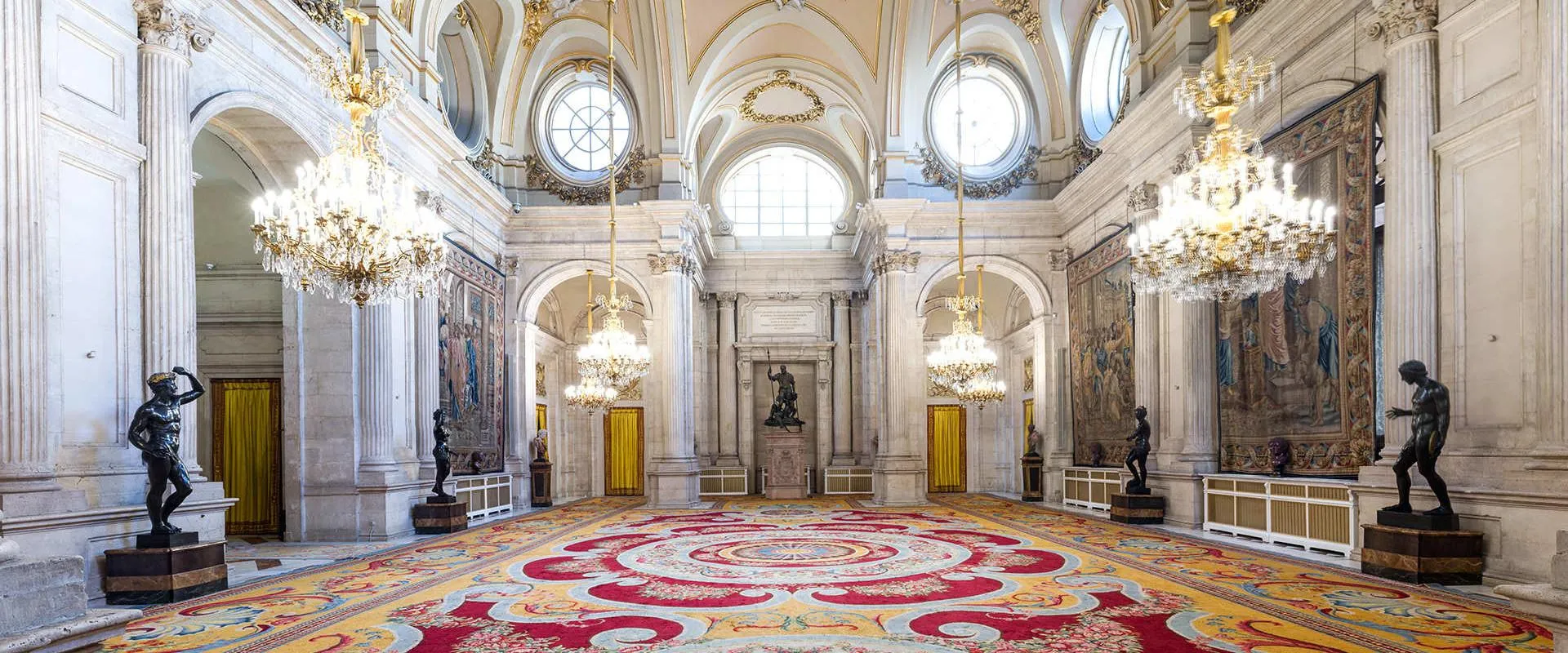 Grote hal met sierlijke kroonluchters, fresco's op het plafond en een groot patroonkleed in het Koninklijk Paleis van Madrid