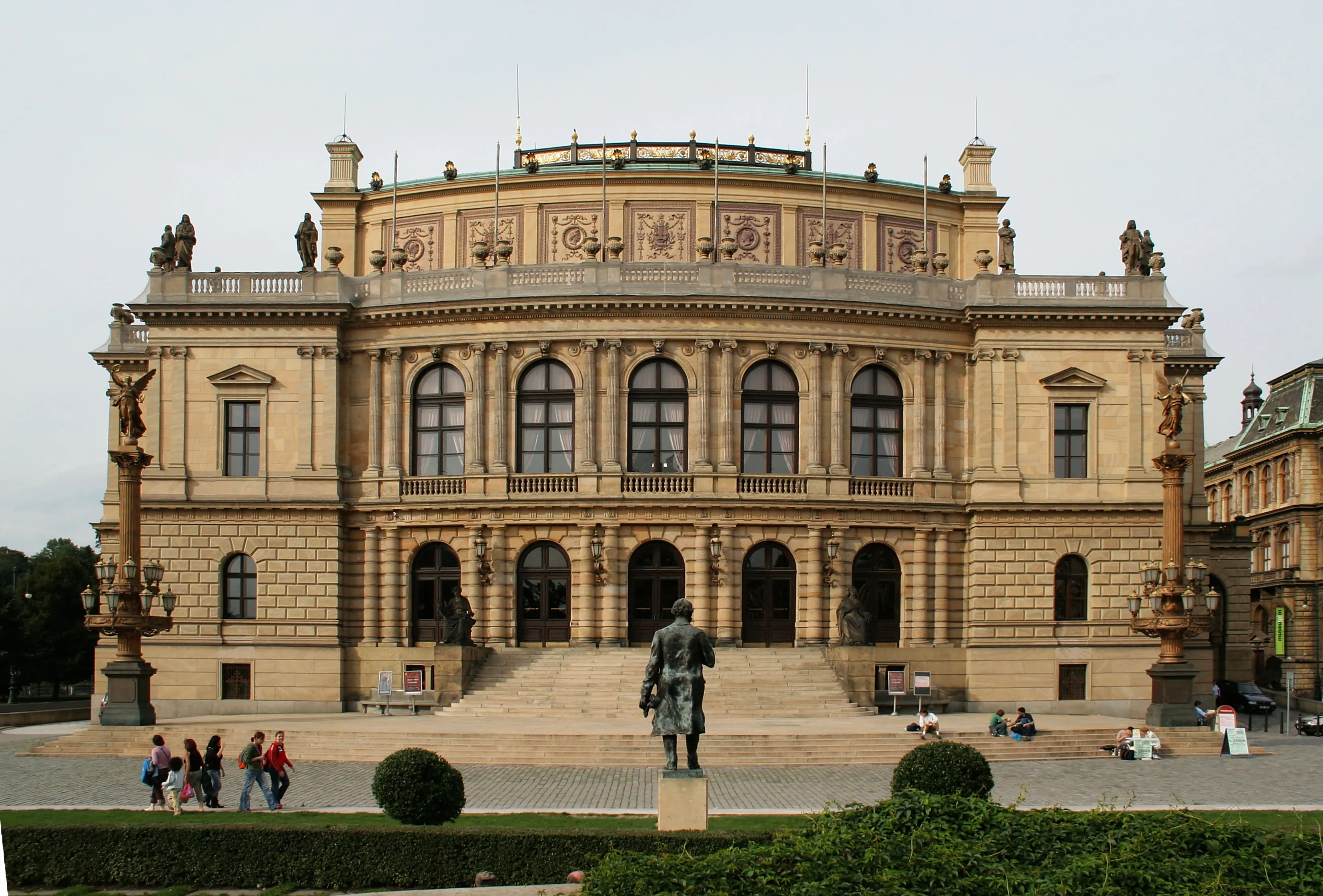 La fachada del Rudolfinum, un gran edificio de piedra con ventanas arqueadas y una escalera central, con gente caminando enfrente.