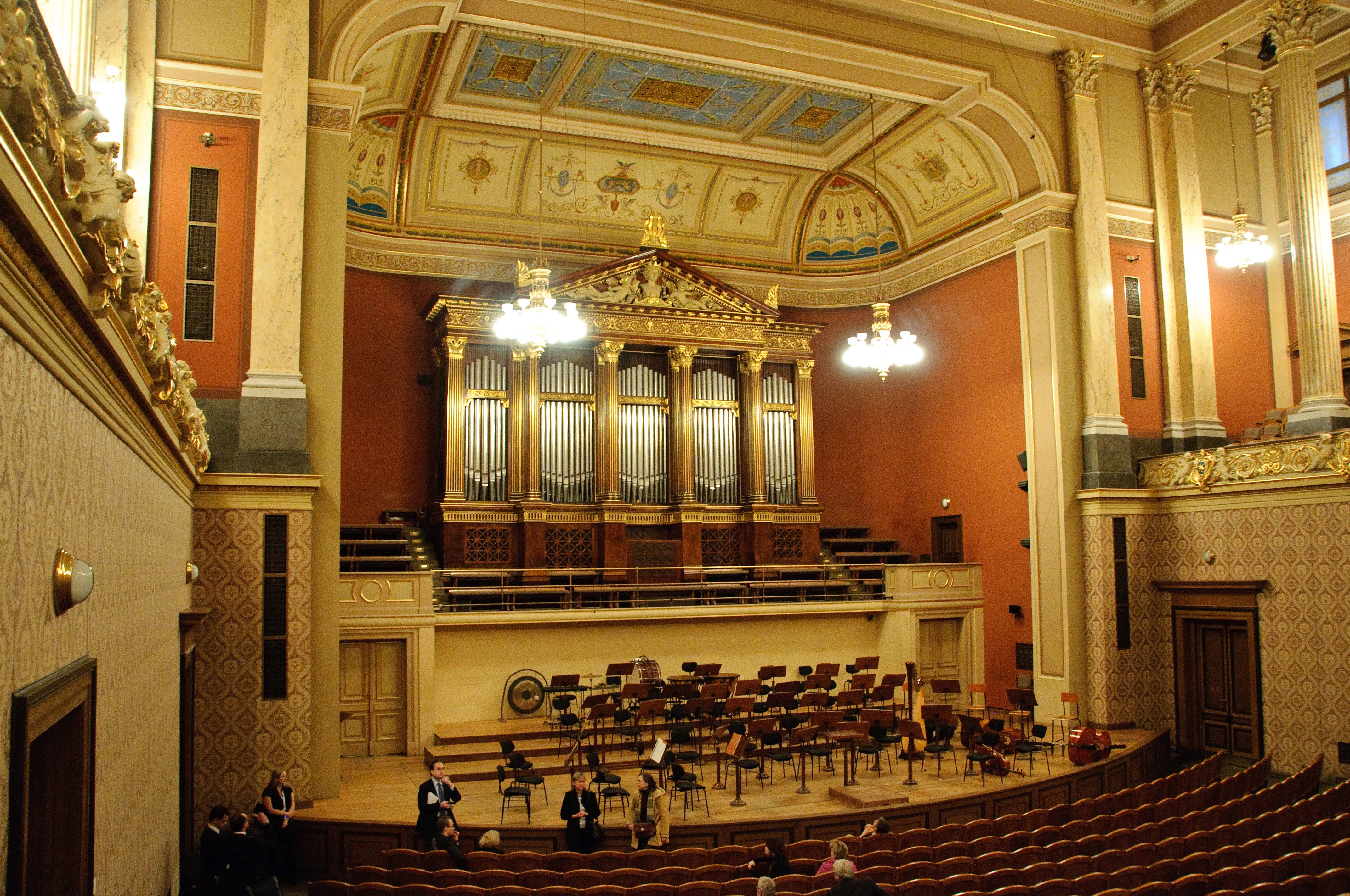 Interior ornamentado de una sala de conciertos con un gran órgano de tubos, decoraciones doradas y filas de asientos bajo un techo decorado