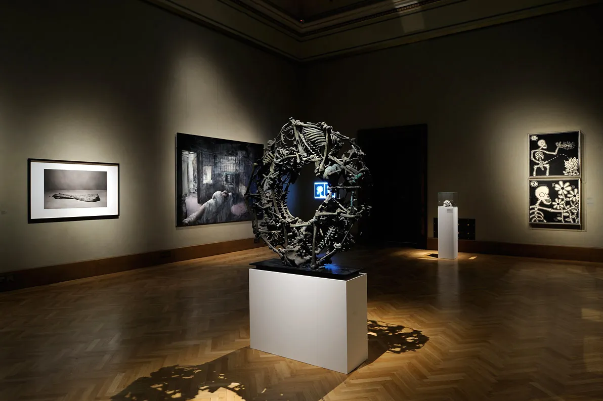 Una gran escultura circular hecha de materiales oscuros sobre un pedestal blanco en una galería, con obras de arte enmarcadas en las paredes y suelos de madera.