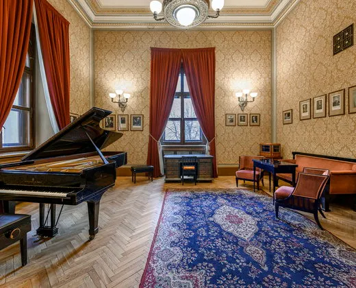 Grand piano in an ornate room with red curtains, patterned wallpaper, and a blue rug