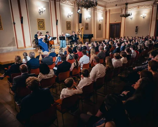 Concert hall with audience seated facing a stage where musicians are performing under chandeliers
