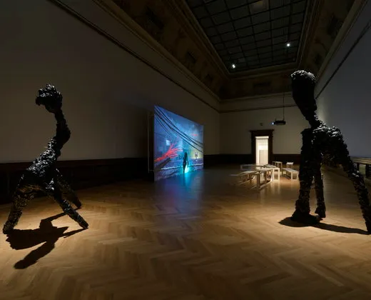 Two dark, textured sculptures in a gallery space with a large screen displaying digital art
