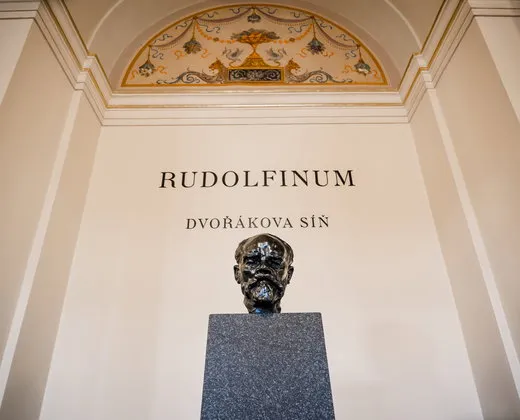 Bust of Antonín Dvořák on a pedestal under an ornate ceiling with 'RUDOLFINUM' and 'DVOŘÁKOVA SÍŇ' written on the wall