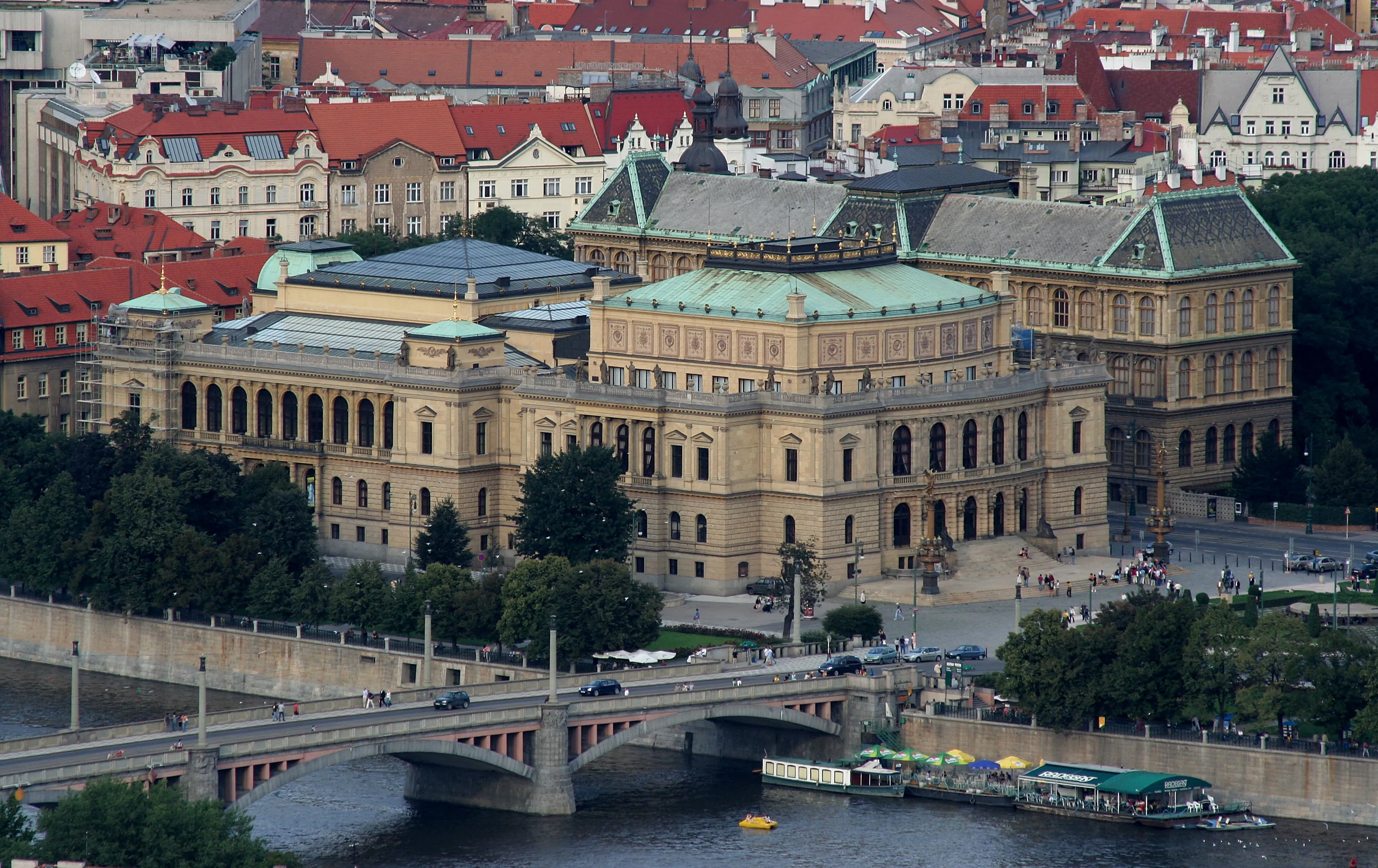 Aerial view of the Rudolfinum concert hall with a green roof, surrounded by red-roofed buildings and a river with a bridge