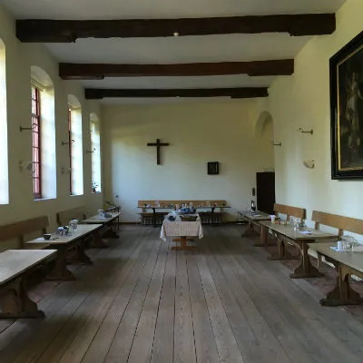 Historische refter met lange houten tafels en banken, zichtbare plafondbalken en een crucifix aan de achterwand.