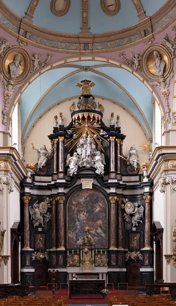 Interior de una ornamentada iglesia barroca que muestra un gran altar mayor con esculturas blancas, columnas de mármol oscuro, detalles dorados y un techo abovedado con medallones pintados y molduras de yeso en tonos rosa y azul